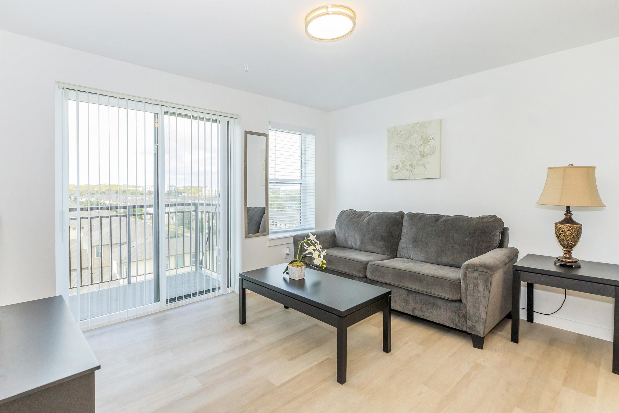 A modern living room featuring a gray sofa, a coffee table, and a lamp. Large windows with blinds allow natural light to enter, and a mirror is mounted on the wall. The decor is minimalistic, with light-colored walls and wooden flooring, creating a spacious and inviting atmosphere.