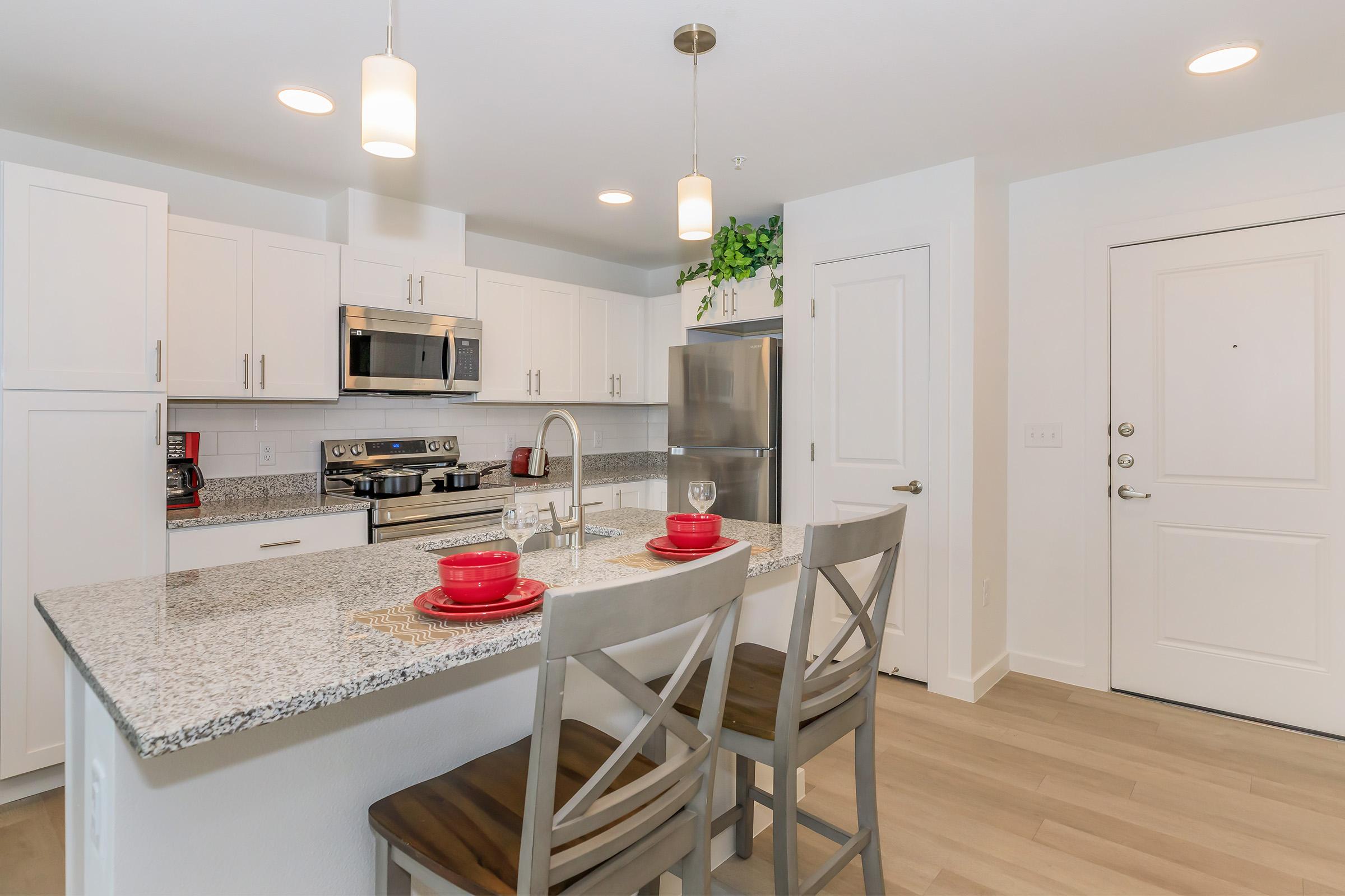 A modern kitchen with white cabinets, stainless steel appliances, and a granite countertop. Two bar stools with beige cushions are positioned at an island, set with red dishes and a wine glass. The space is well-lit with pendant lamps, and there are green plants and a door visible in the background.