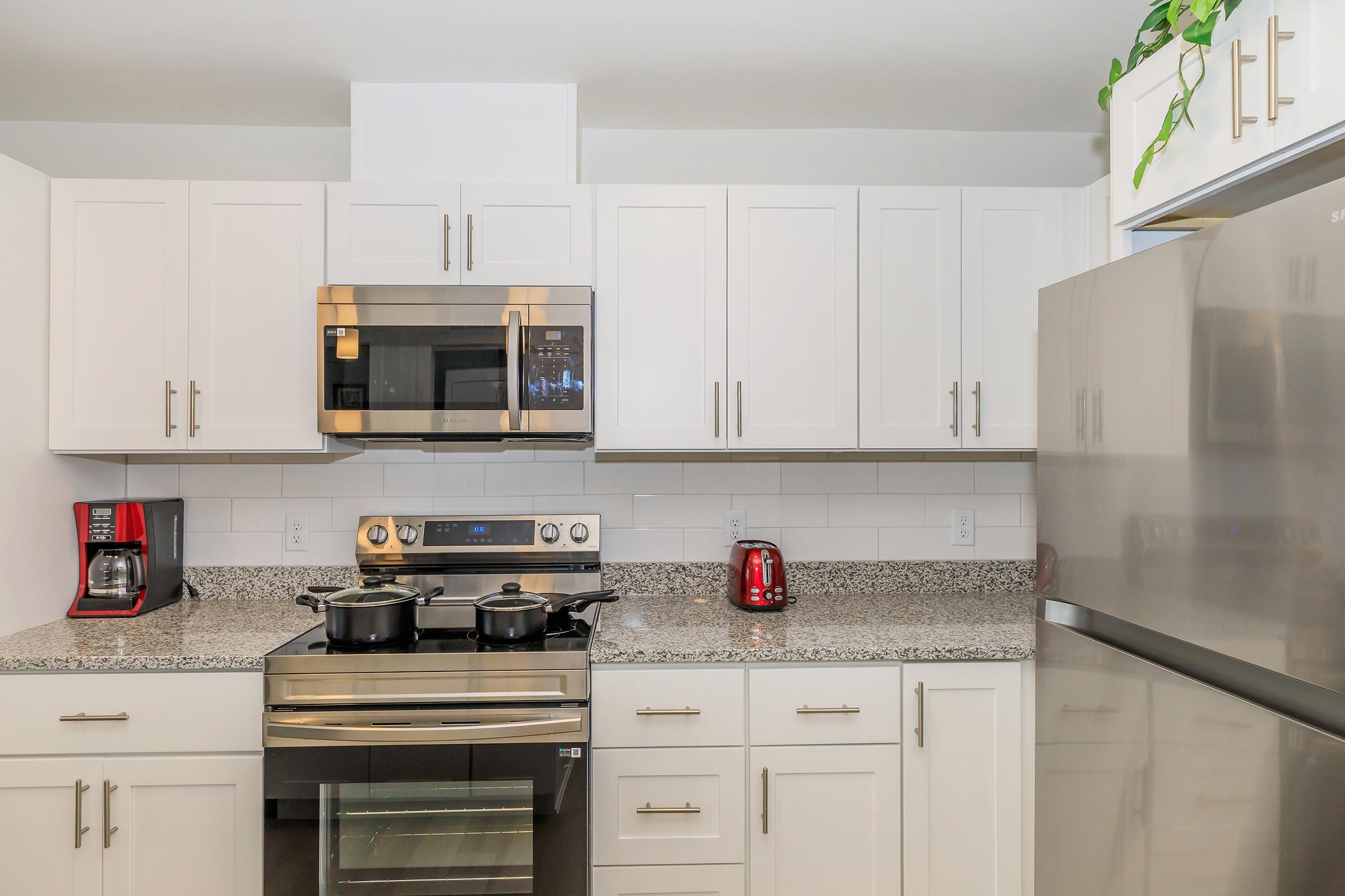 Modern kitchen featuring white cabinetry, stainless steel appliances, and a gray granite countertop. Includes an electric stove, microwave, and coffee maker, along with a red kettle, and decorative plant on top of the cabinets. Bright and inviting atmosphere with ample storage space.