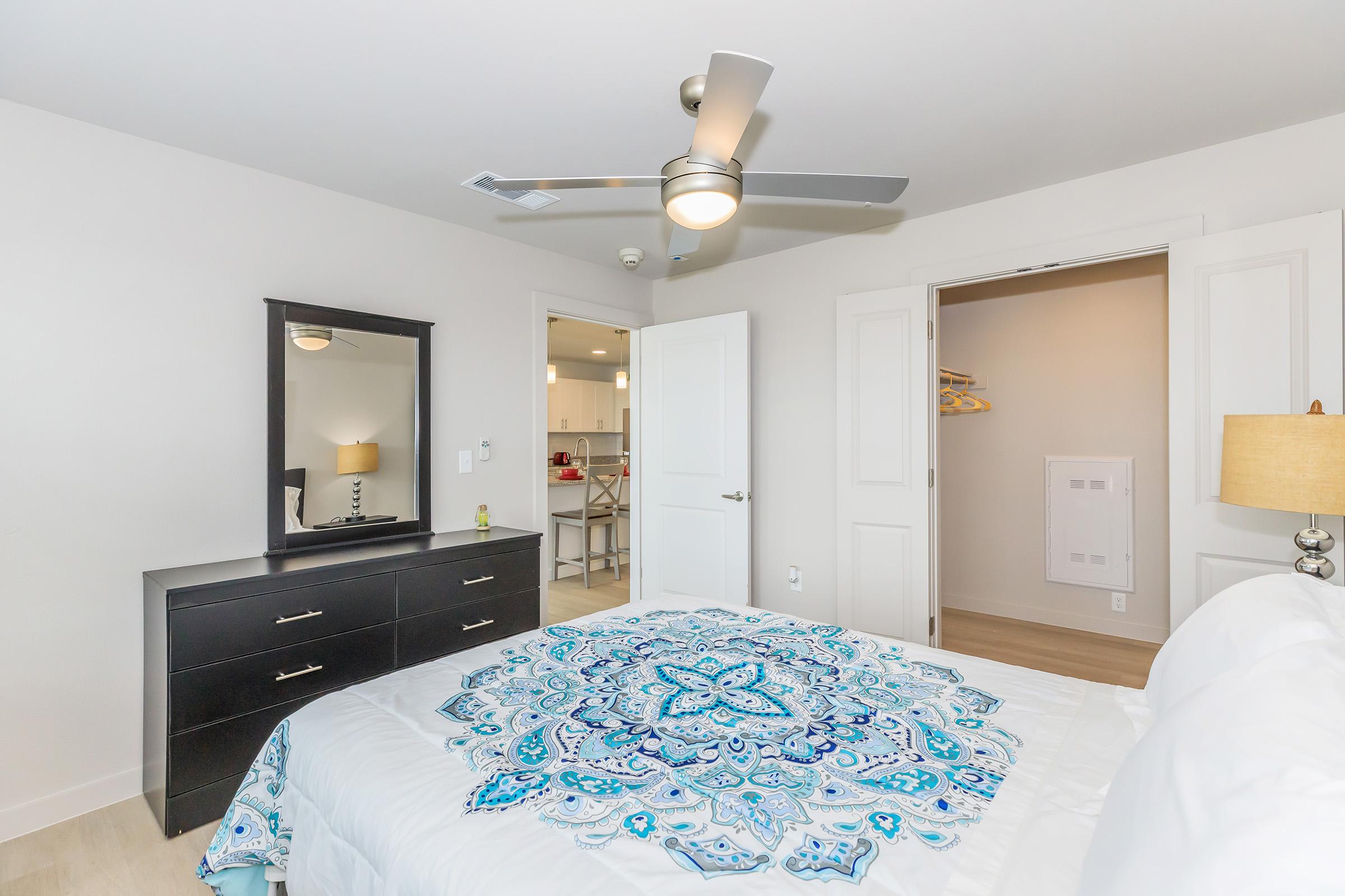 Modern bedroom featuring a neatly made bed with a vibrant blue and white patterned duvet. A sleek black dresser with a mirror is positioned alongside the bed. The room has white walls, a ceiling fan, and an open doorway leading to a closet or additional space, providing a bright and airy atmosphere.