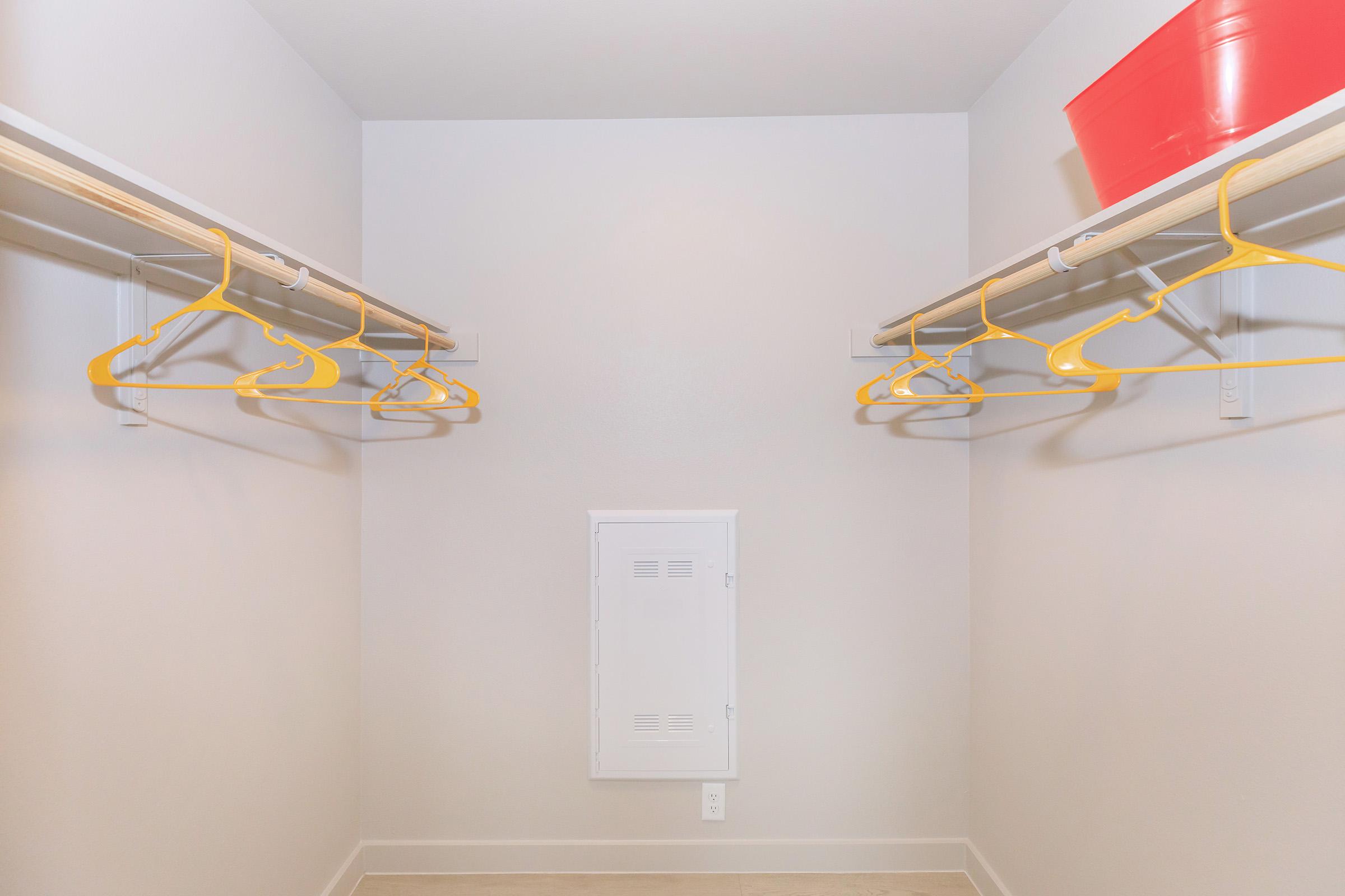 A spacious, empty closet featuring two rows of yellow hangers on wooden rods, with a small white panel on the wall and a red storage box on the top shelf. The walls are light-colored, and the floor is neutral-toned, creating a clean and open appearance.