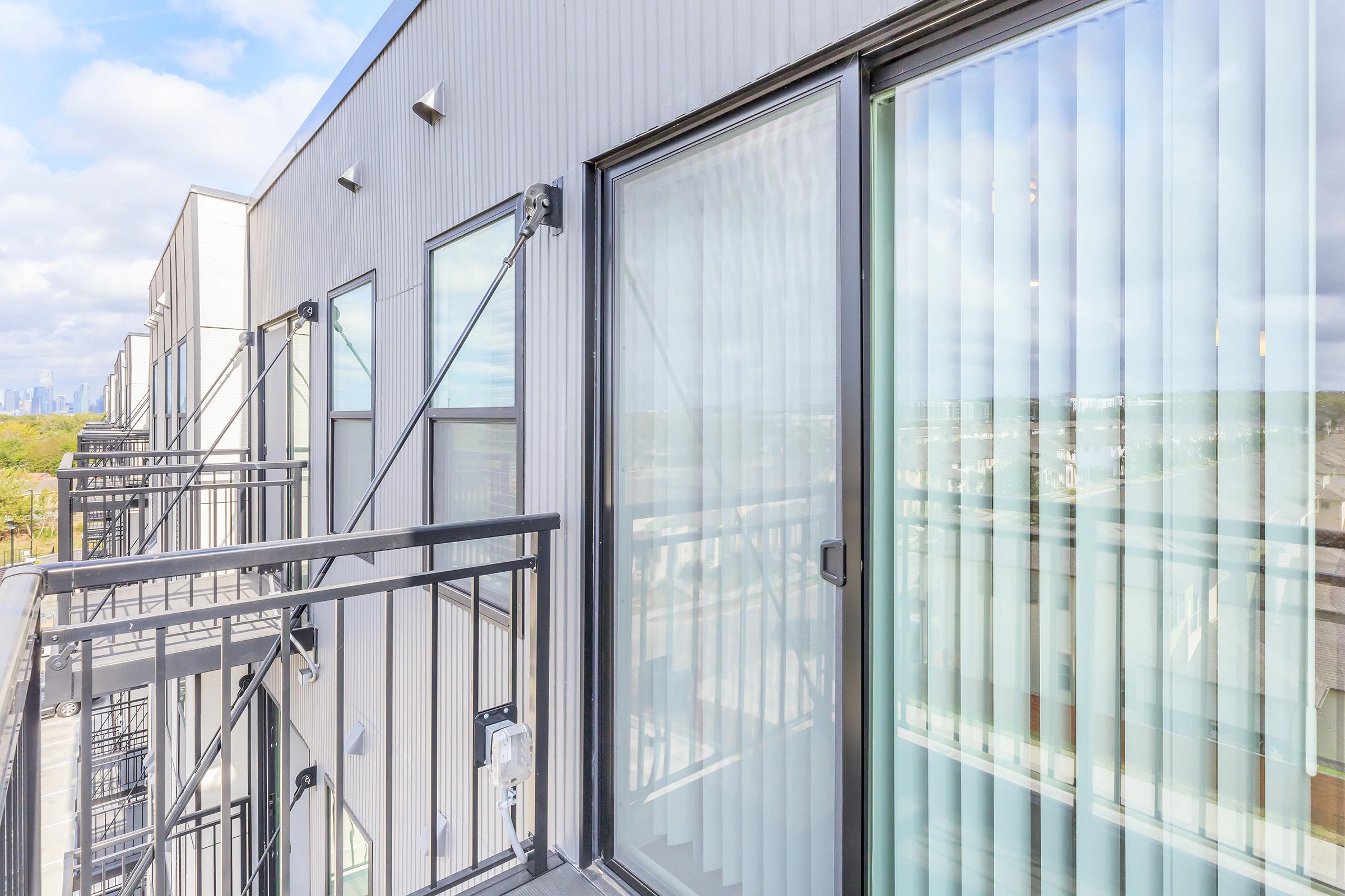 Exterior view of a modern apartment building featuring multiple balconies with sliding glass doors. The balconies are equipped with railings, and the building has a sleek design with large windows. In the background, there are hints of a city skyline visible on a clear day.