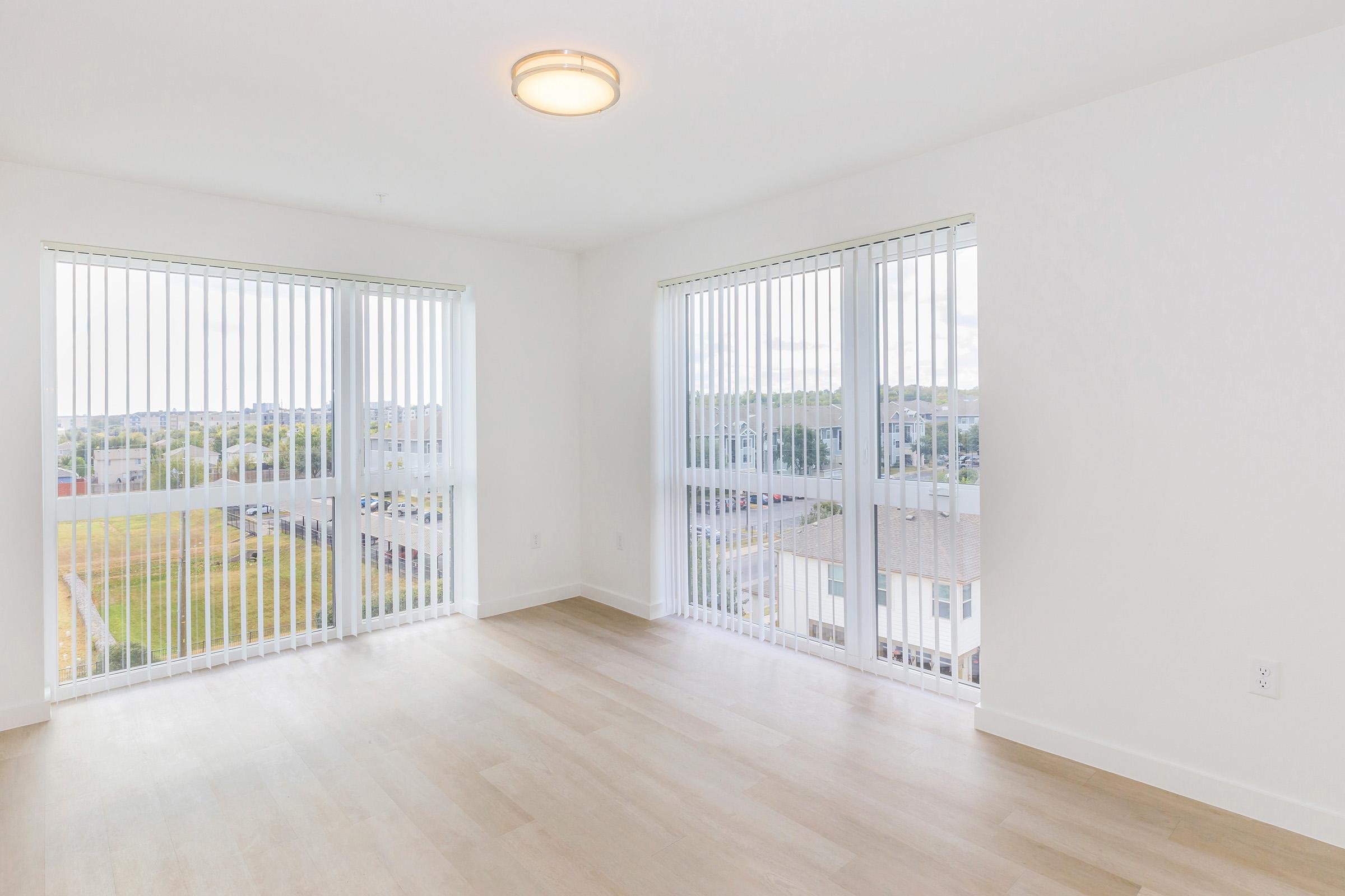 Bright and spacious empty room with large vertical blinds covering two windows, allowing natural light to stream in. The floor is light wood, and the walls are painted white. The view outside features greenery and buildings. A modern ceiling light fixture is visible.