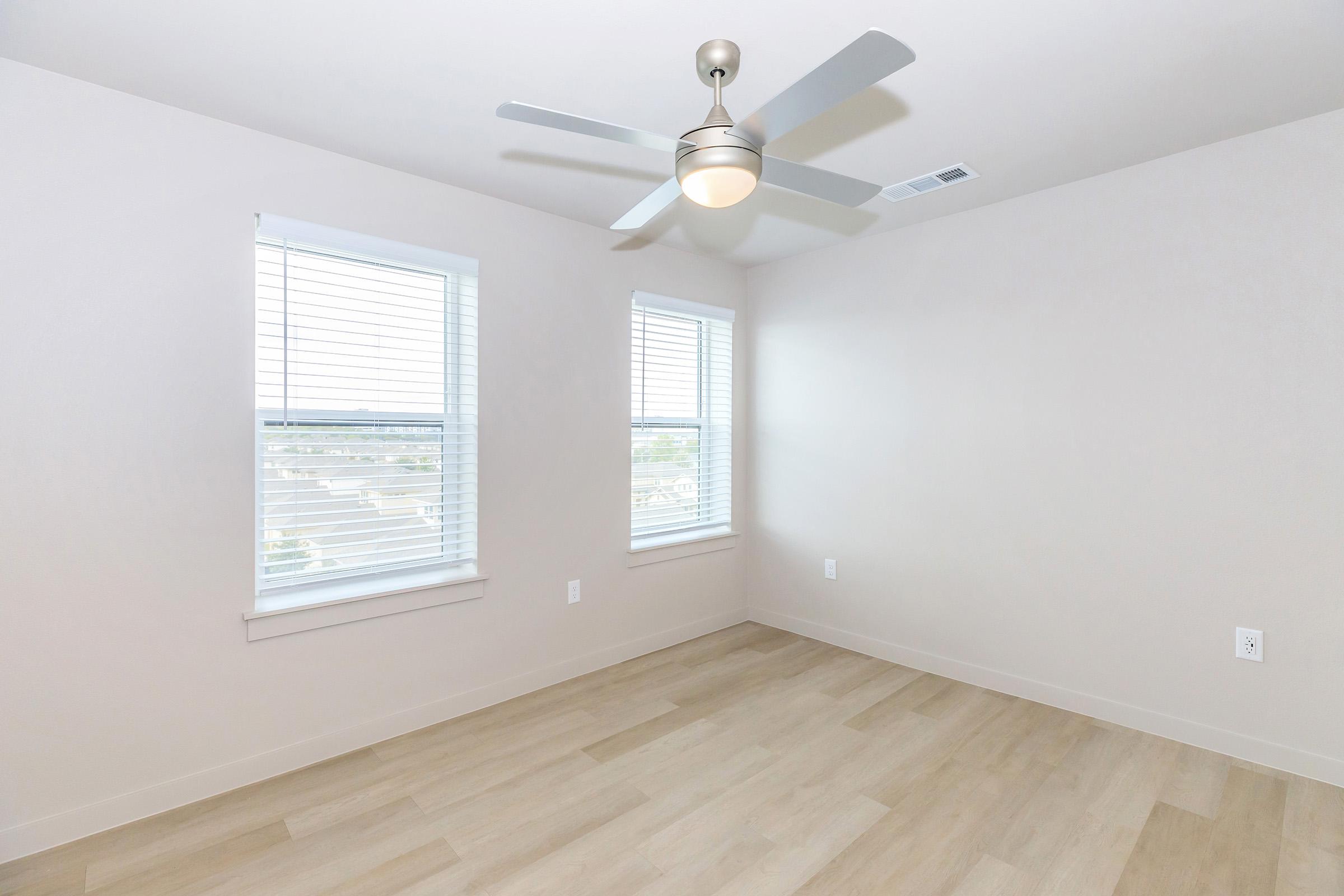 A clean, empty room featuring light-colored walls, a ceiling fan, and two windows with blinds. The floor is light wood laminate, creating a bright and airy atmosphere. No furniture is present, emphasizing the spaciousness of the area.