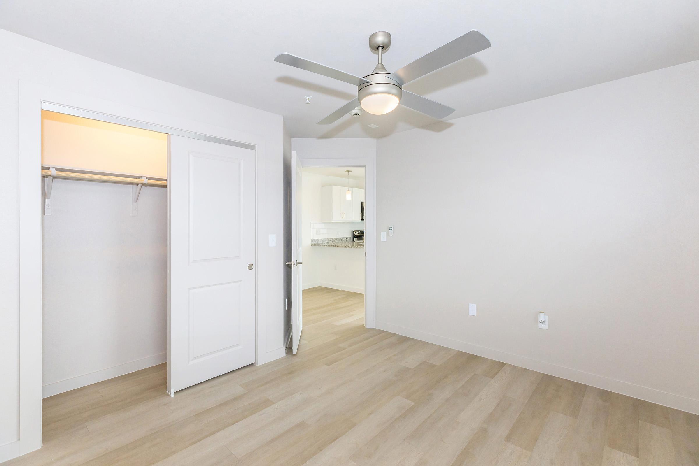 A spacious, empty room featuring light-colored walls and wooden flooring. There's a ceiling fan in the center, and an open closet with sliding doors on the left. A door to another room is visible in the background, suggesting access to a kitchenette or bathroom area. The overall ambiance is modern and clean.