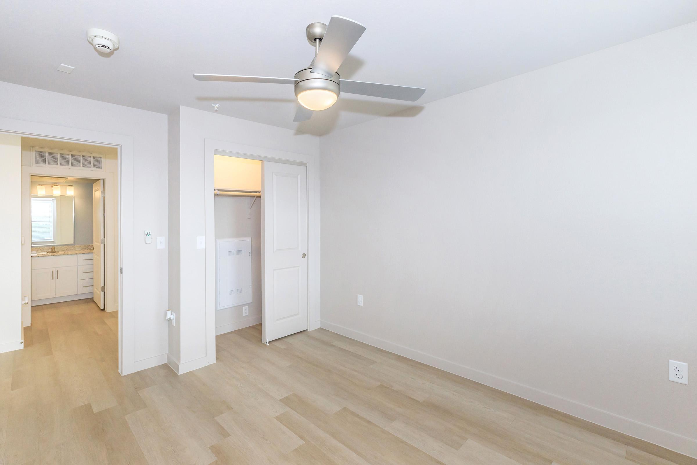 Bright, empty room with light-colored walls and wooden flooring. A ceiling fan is installed, and there is a doorway leading to a bathroom area with a mirror and cabinetry visible through an open door. The space is well-lit, offering a modern and clean aesthetic.