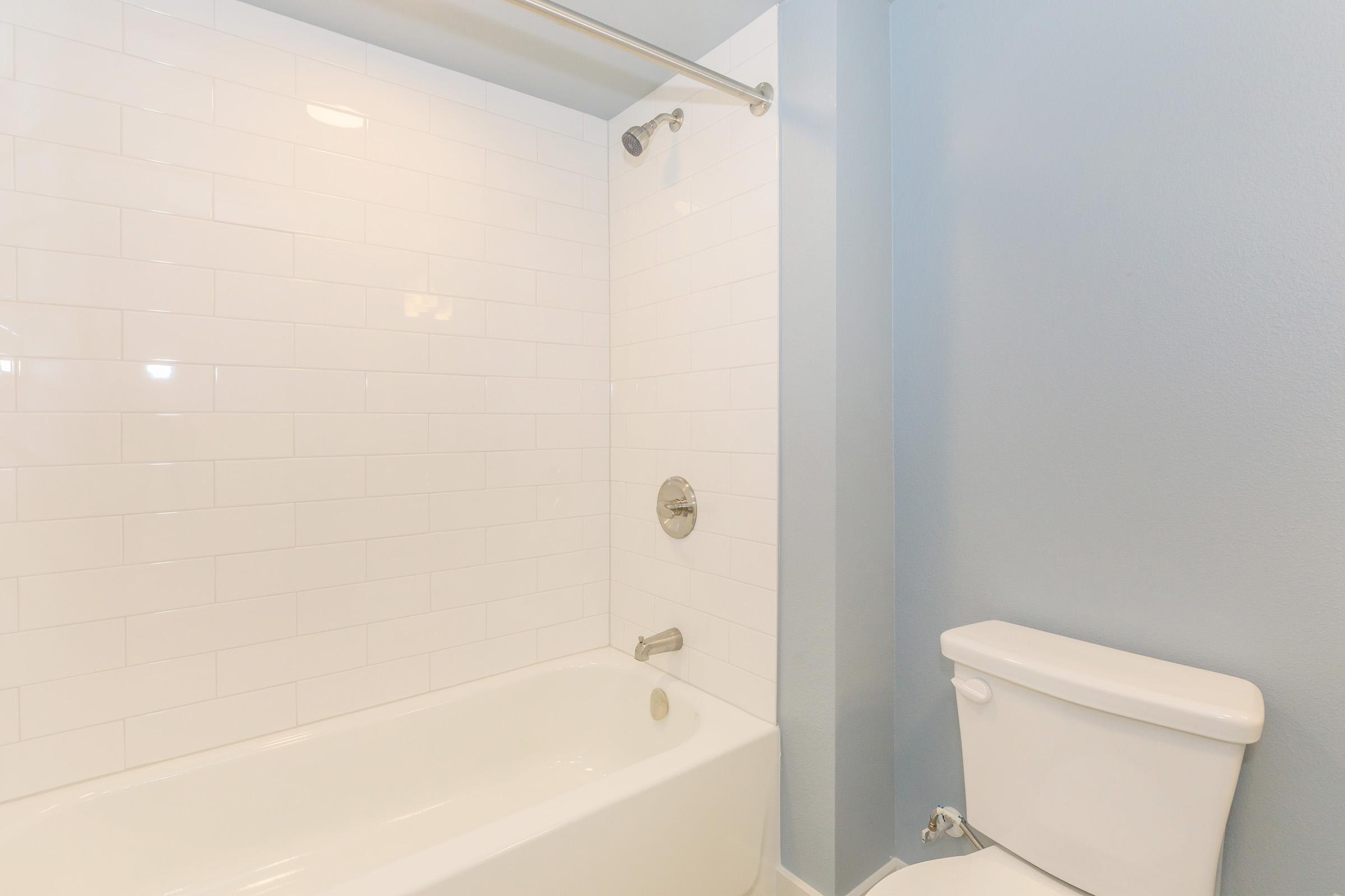 A clean, modern bathroom featuring a white tile shower with a glass door and a bathtub. Adjacent to the bathtub is a white toilet against a soft blue wall, enhancing the room's fresh and contemporary aesthetic. The lighting is bright, creating an inviting atmosphere.