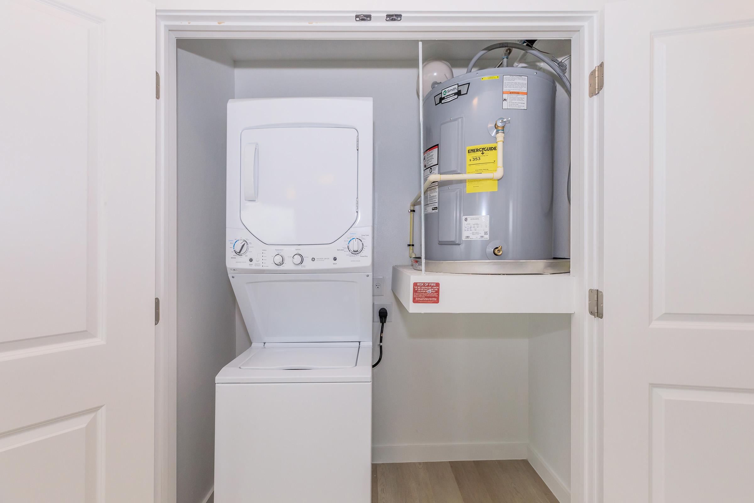 A compact laundry area featuring a stacked washer and dryer on the left, with a water heater mounted on a shelf to the right. The space has white doors and light wood flooring, creating a clean and organized appearance.