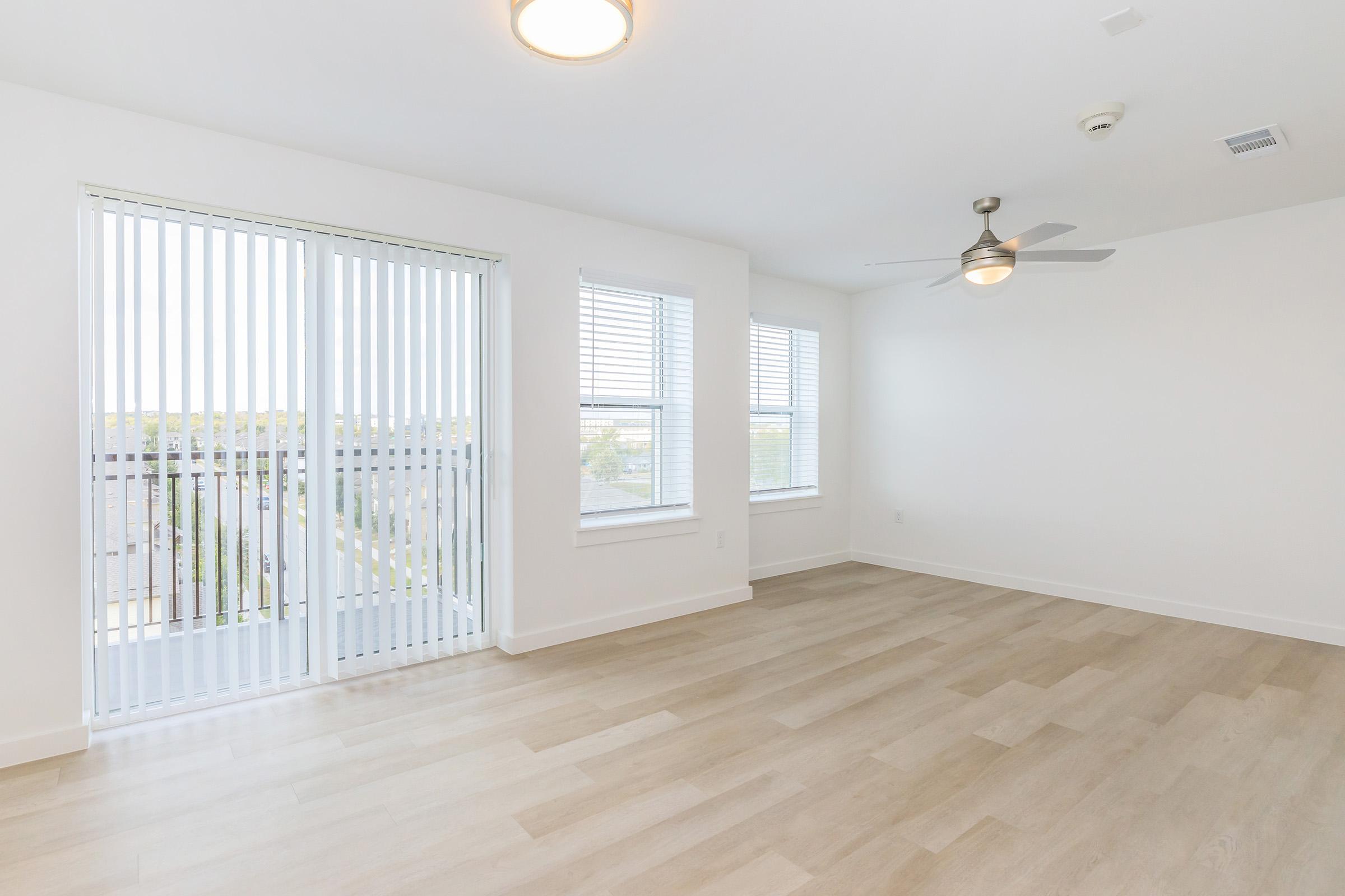 A spacious, empty room with light wood flooring, two large windows with white blinds, and a ceiling fan. The walls are painted white, and there is a modern light fixture on the ceiling. The room has a clean and bright feel, ideal for various purposes.