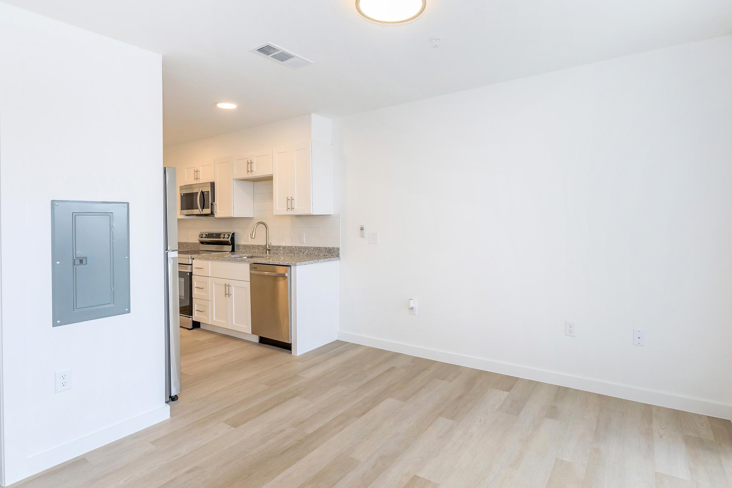Modern kitchen in an open-concept living space, featuring white cabinetry, stainless steel appliances, a granite countertop, and light-colored hardwood flooring. The room is well-lit, with a blank wall on the right side and a utility panel on the left.