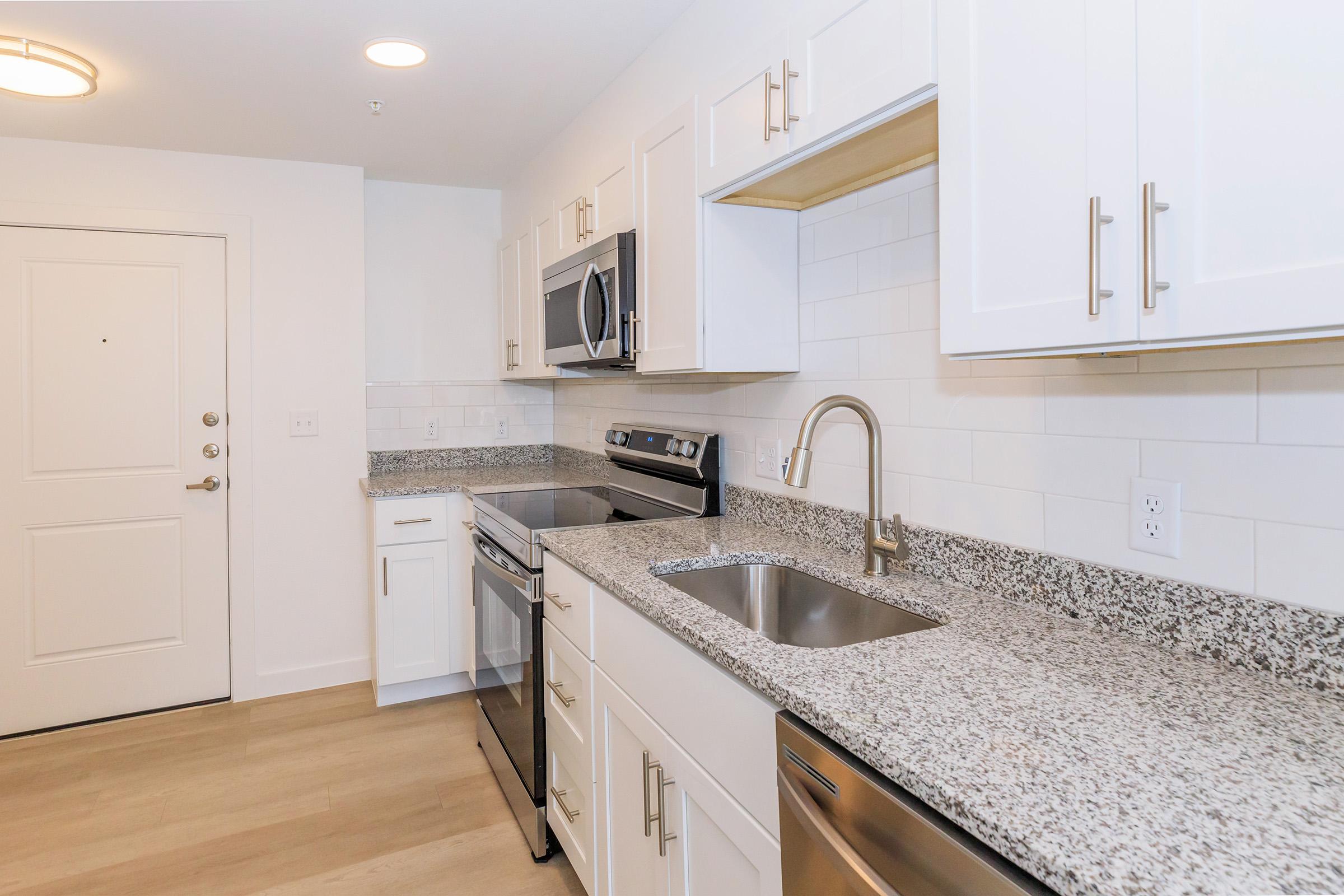Modern kitchen featuring white cabinetry, stainless steel appliances including a microwave and stove, a single-basin sink, and a granite countertop. The space has a clean, contemporary look with light-colored wood flooring and white backsplash tiles. A door leads to the exterior.