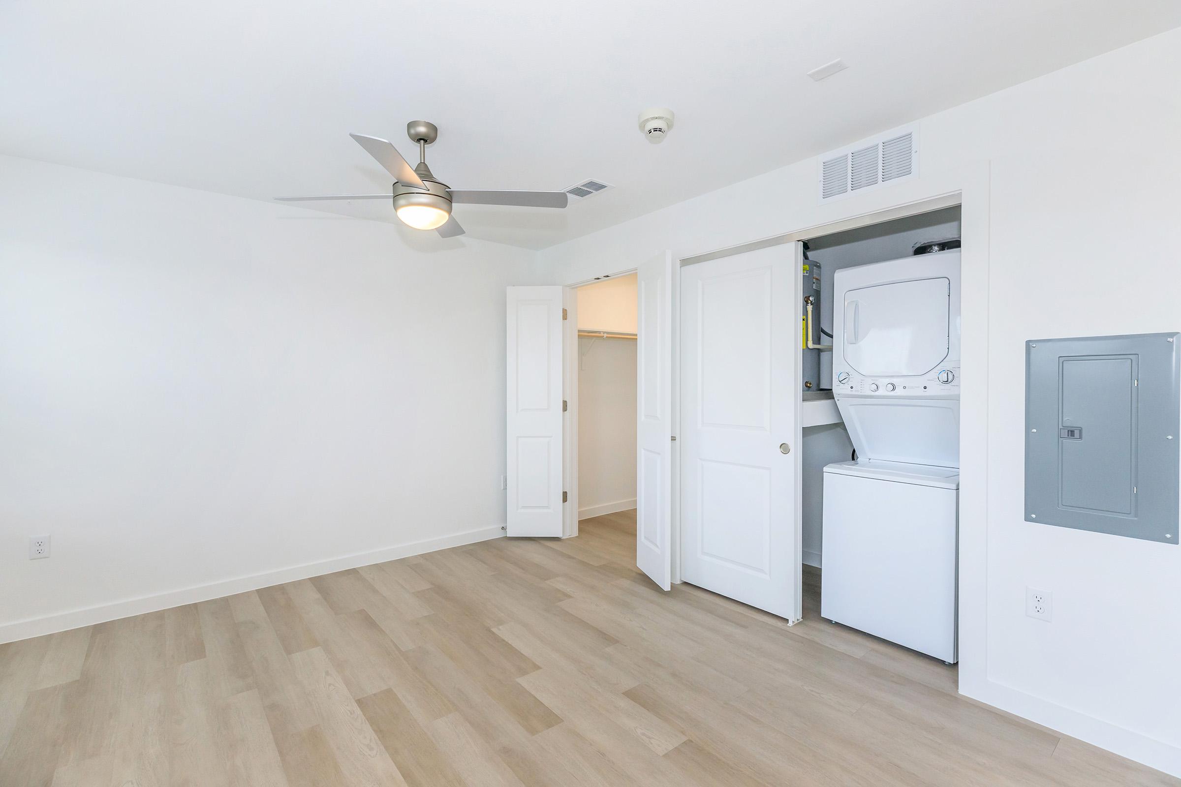 A bright and modern room with light-colored walls and wooden flooring. It features a ceiling fan, a closet with double doors, and a stacked washer and dryer unit. The space is well-lit, creating an inviting atmosphere.