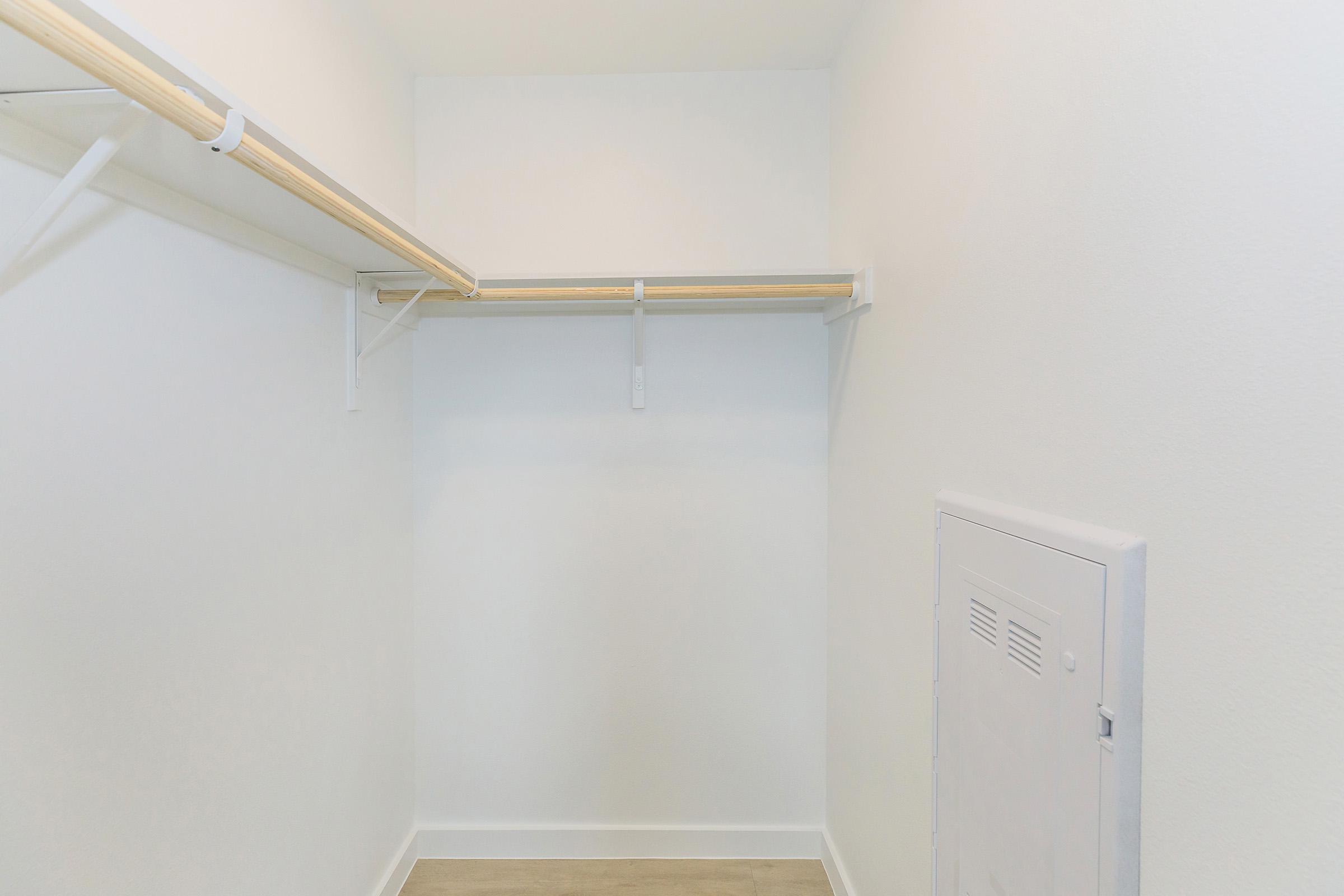 Empty closet with white walls and light wood flooring. There is a single wooden rod for hanging clothes installed on the left side, and a small utility panel on the right wall. The space is bright and uncluttered, creating an open feel.
