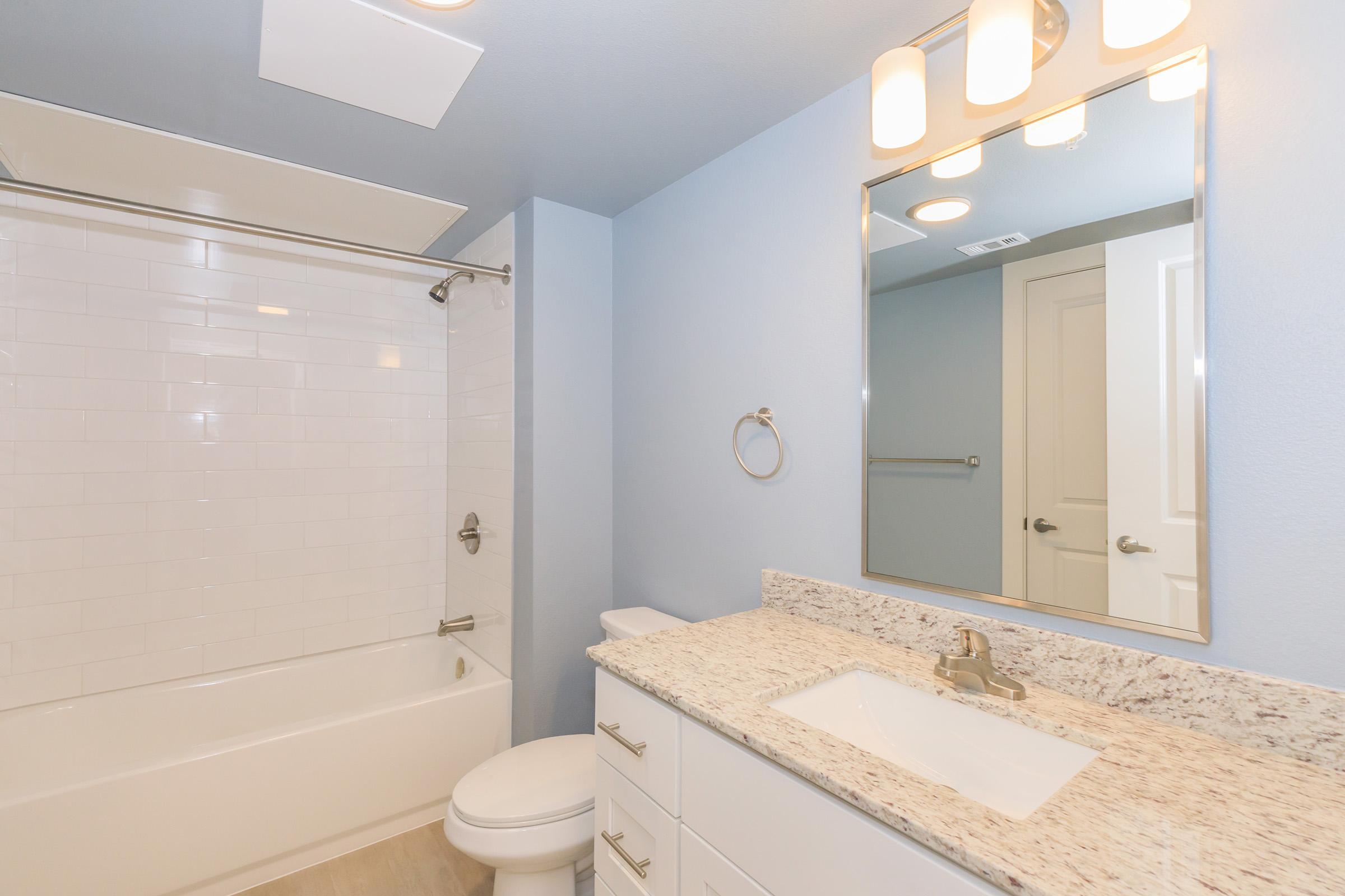 A modern bathroom featuring a bathtub with a shower curtain, a white sink with a granite countertop, a mirror above the sink, and light fixtures. The walls are painted in a soft blue, and there are white tiles and a door visible in the background.