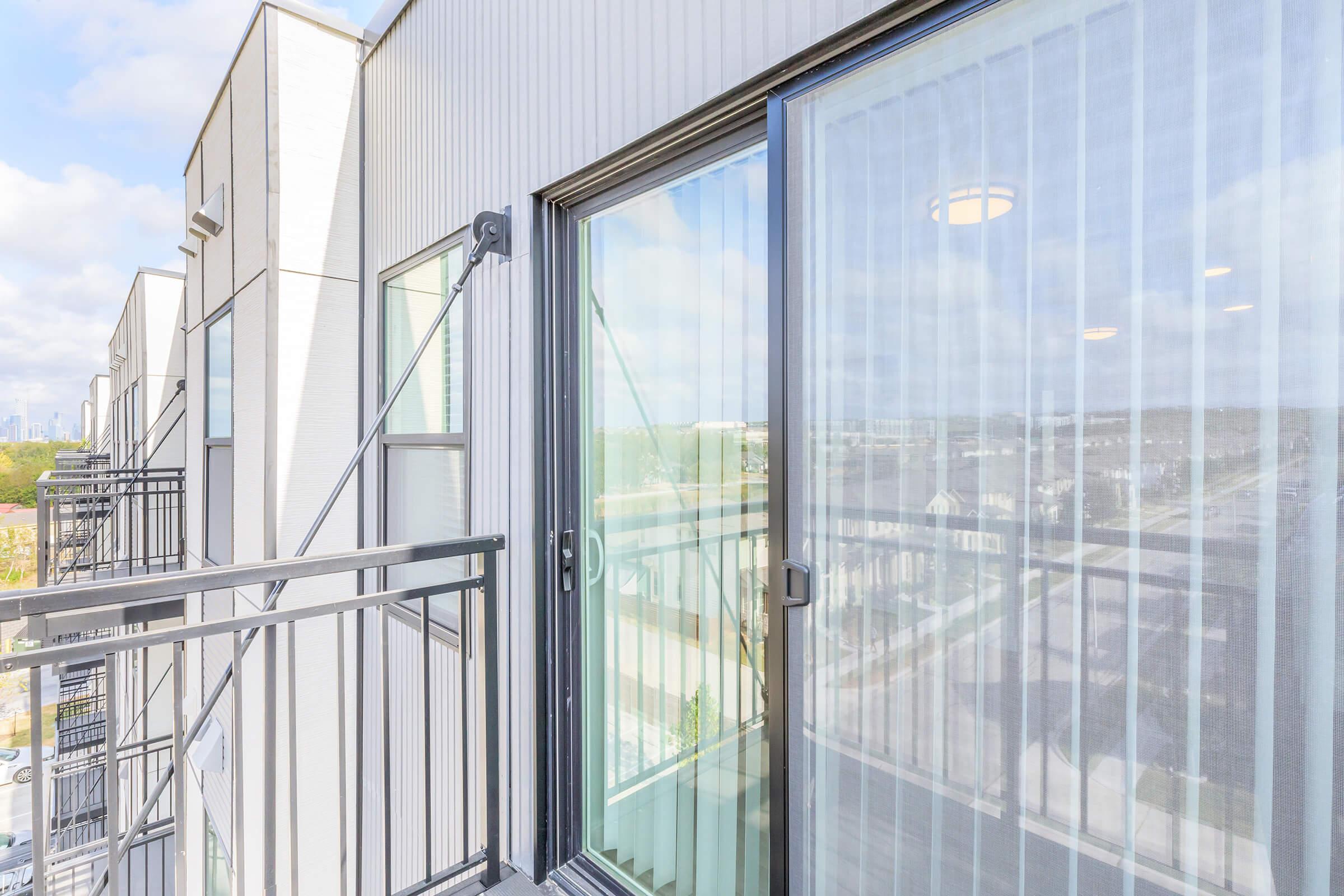 A modern balcony with sliding glass doors, overlooking a scenic urban view. The balcony features metal railings and is designed with clean lines, showcasing a bright and airy atmosphere with natural light filtering through sheer curtains.