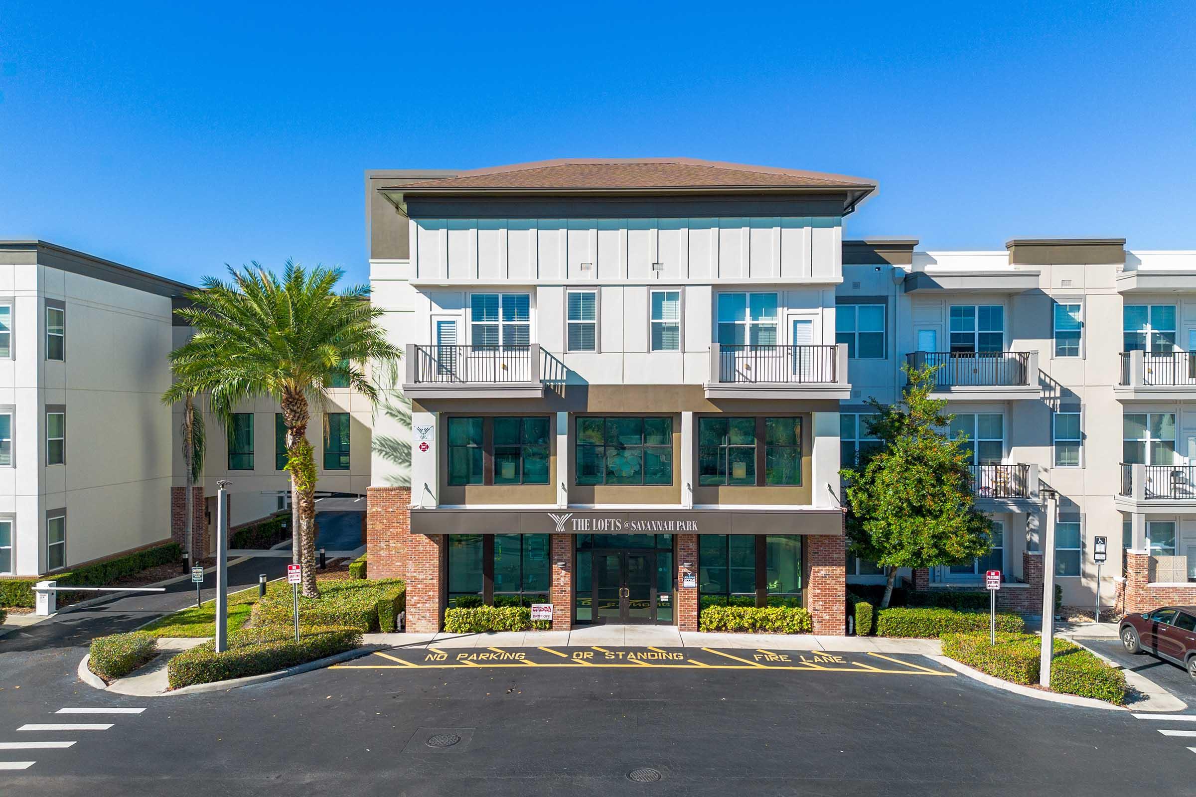 A modern multi-story building with a flat roof, featuring large windows and balconies. The entrance is framed by palm trees and well-maintained greenery. The pavement in front displays "No Parking" and "Car Stationing" signs. A clear blue sky provides a bright backdrop.