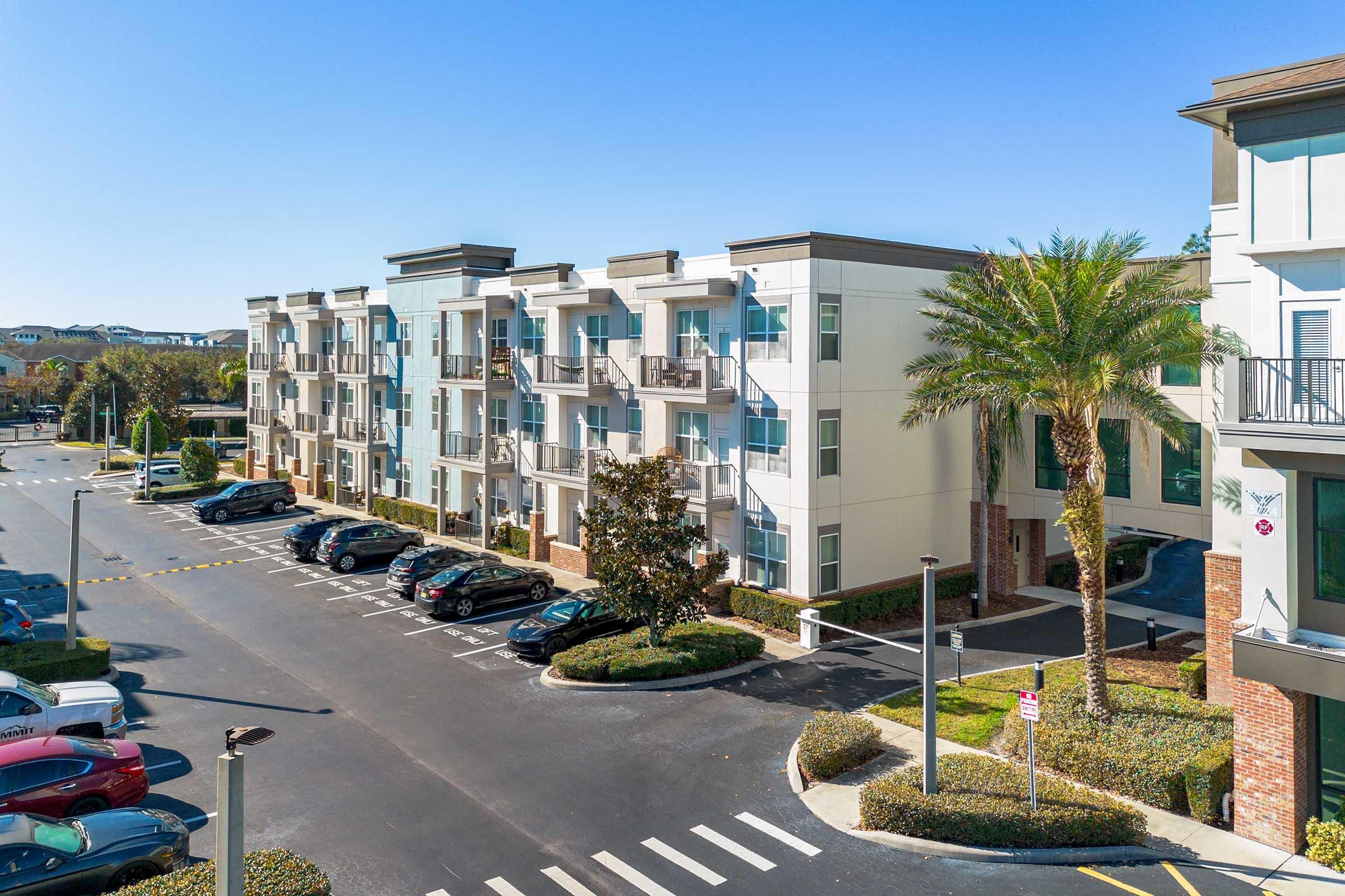 Modern apartment community with several three-story buildings, featuring balconies and large windows. There is a parking lot with parked cars, palm trees, and neatly landscaped greenery. The sky is clear and bright. The scene is well-lit, showcasing a sunny day in an urban environment.