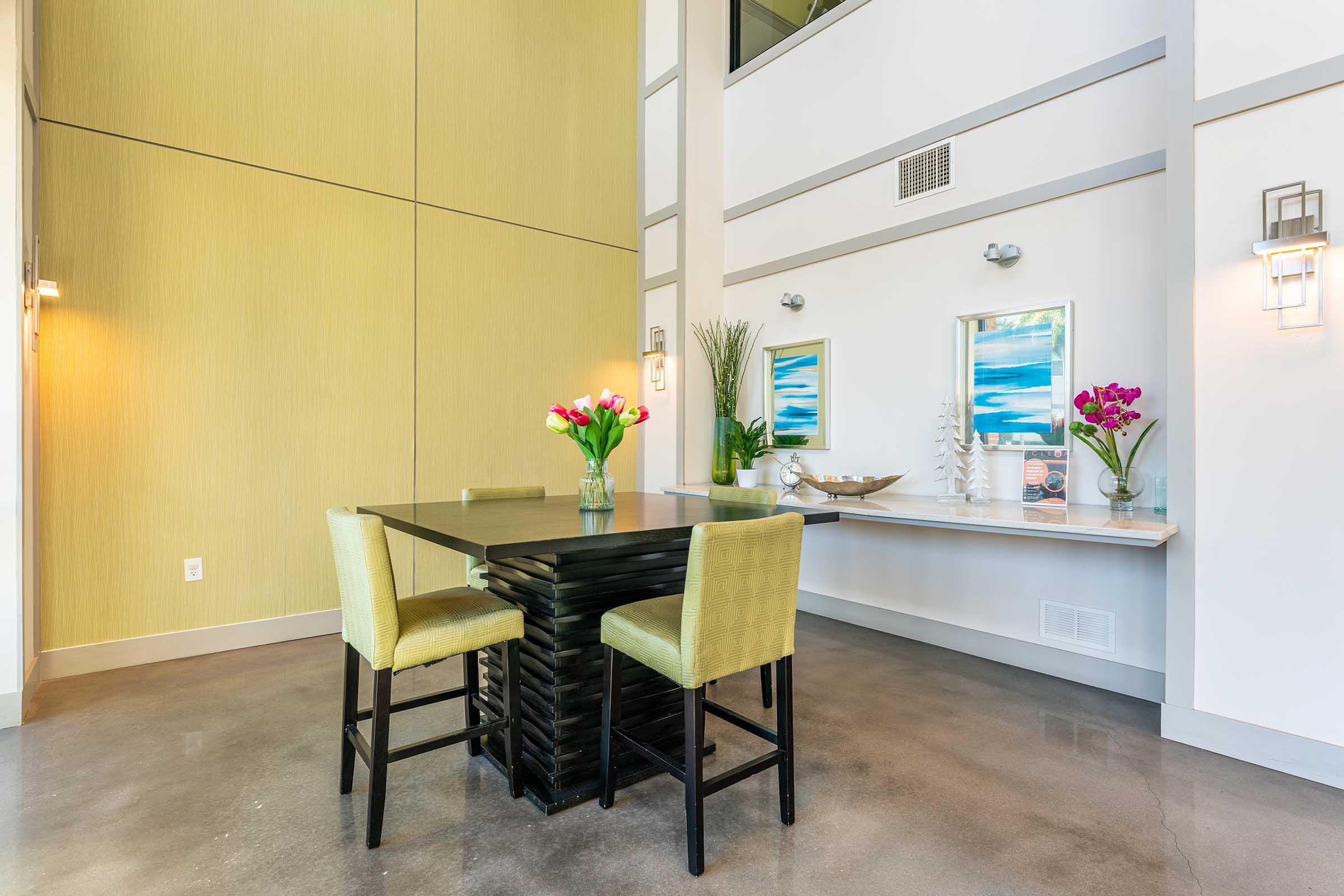 A modern interior with a small table and two green chairs. There are fresh flowers in a vase on the table. The walls feature light-colored panels and artwork, with decorative items on a ledge and plants adding a touch of nature. The floor is polished concrete, creating a sleek and contemporary look.