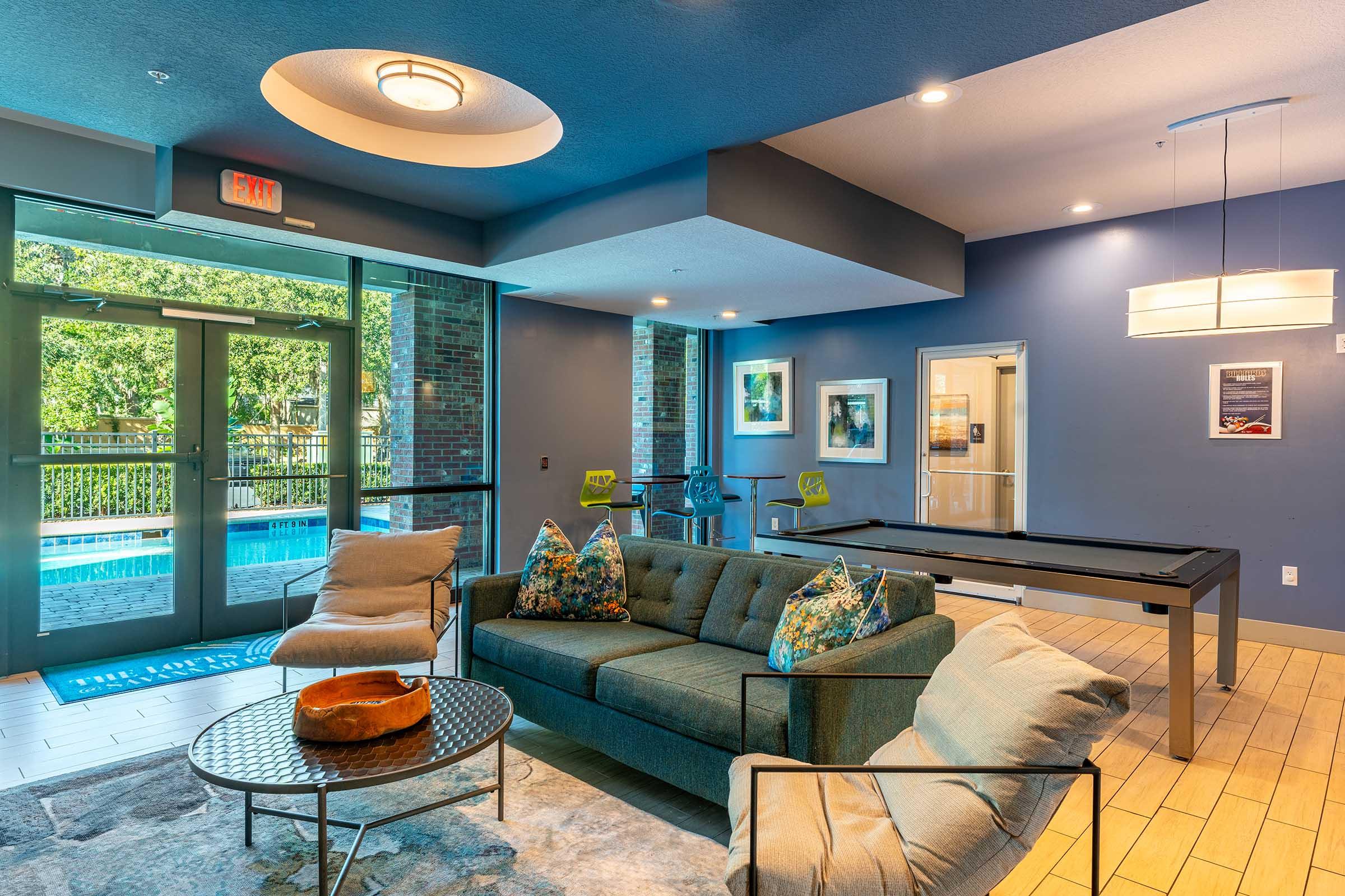 A modern lounge area featuring a comfortable gray sofa with decorative pillows, a round coffee table, and a pool table. Large windows provide natural light, offering a view of the outdoor pool. The walls are painted in a calming blue, and stylish light fixtures add a contemporary touch.