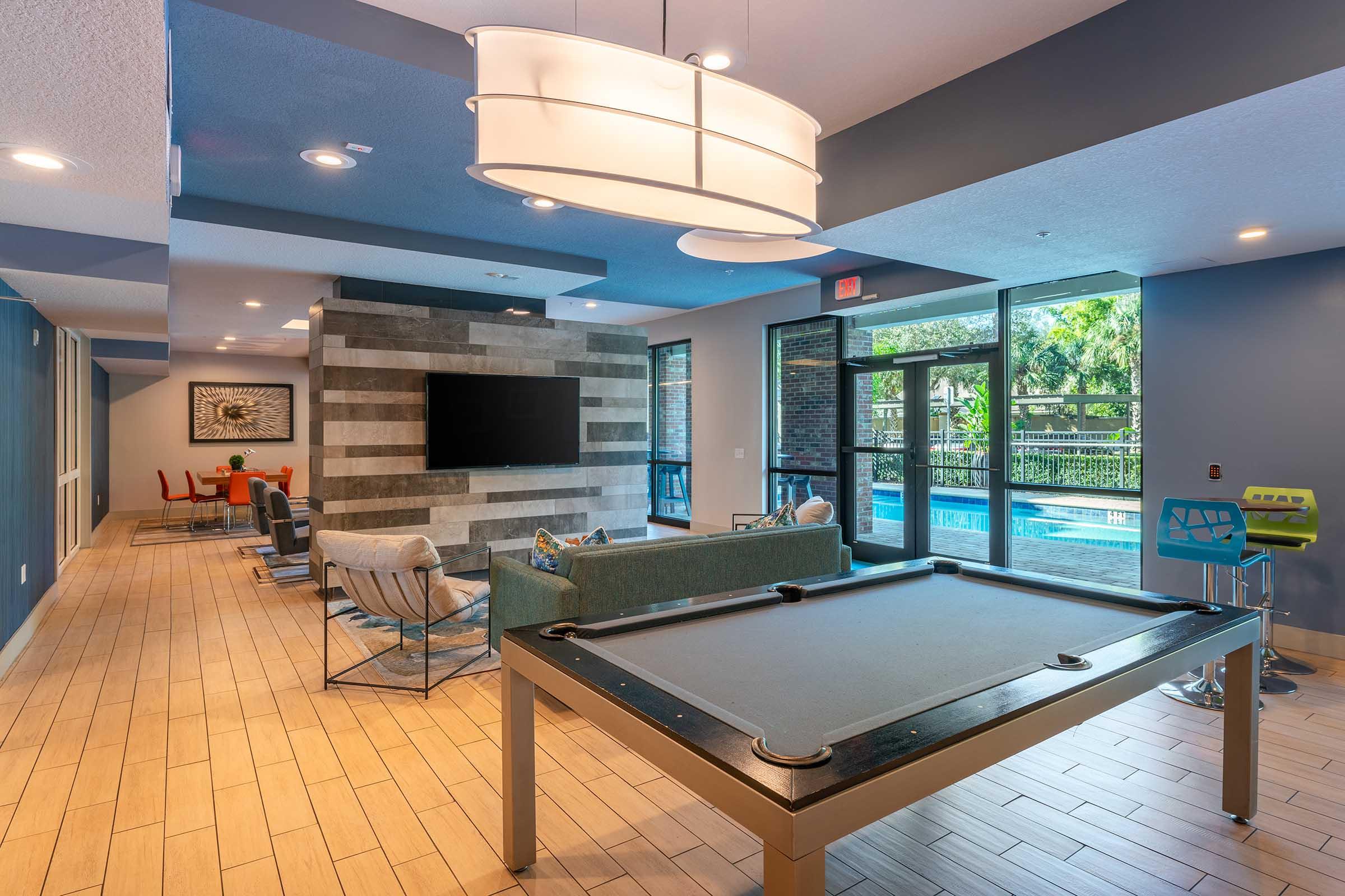 A modern lounge area featuring a pool table, comfortable seating, and a large flat-screen TV. Large windows provide views of an outdoor pool area. The decor includes a mix of blue and gray tones, with stylish lighting and contemporary furniture, creating a welcoming and inviting atmosphere.