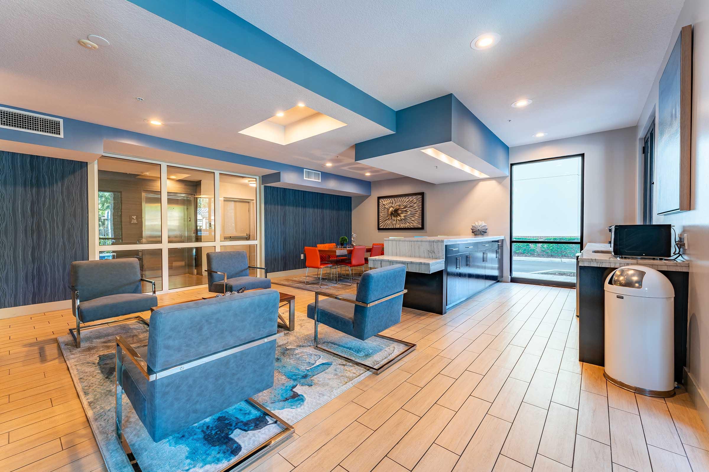 A modern lounge area featuring comfortable seating with gray chairs and a red dining set. There is a stylish kitchenette with a countertop, and large windows allow natural light to fill the space. The floor is made of light wood tiles, and decorative artwork adorns the walls.