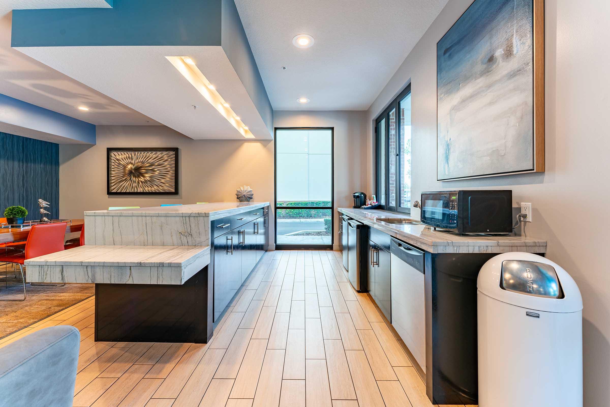 Modern kitchen interior featuring a sleek design with a marble countertop island, black cabinetry, and stainless steel appliances. Large windows bring in natural light, offering a view of the outdoors. The space is stylishly decorated, including a contemporary artwork piece on the wall.