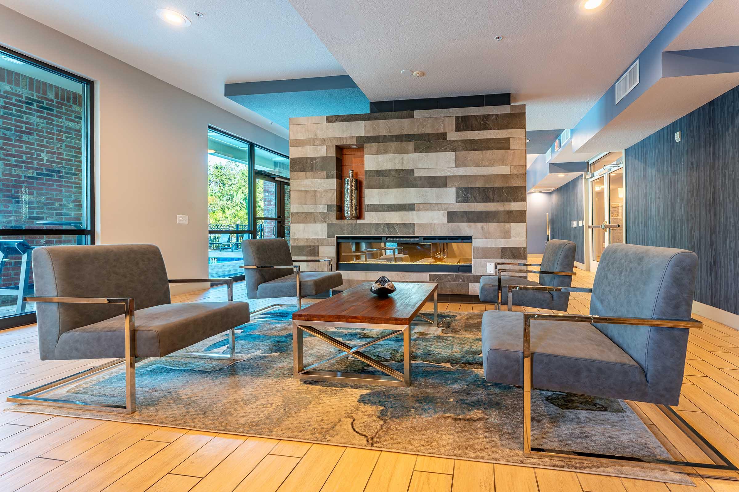 A modern lounge area featuring four gray chairs arranged around a wooden coffee table on a decorative rug. The backdrop includes a stone fireplace and large windows with natural light illuminating the space, creating a welcoming atmosphere.