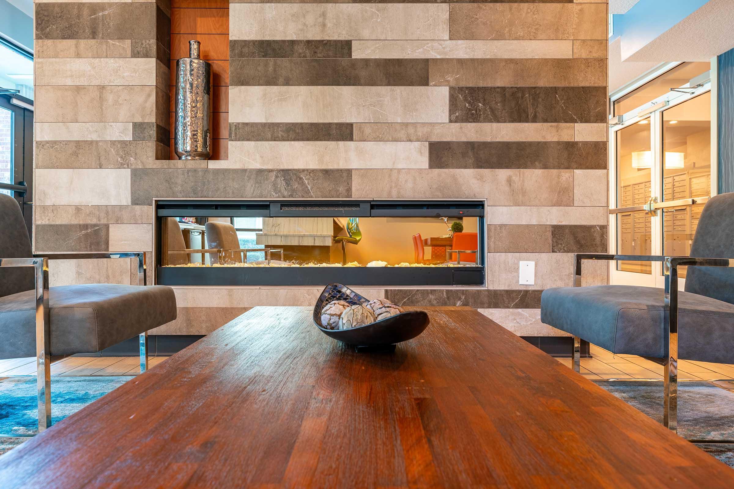 Modern interior featuring a stylish fireplace set into a stone wall, flanked by two gray armchairs. A wooden coffee table with a decorative bowl sits in the foreground, enhancing the contemporary aesthetic of the space. Bright light floods the area, creating a warm inviting atmosphere.