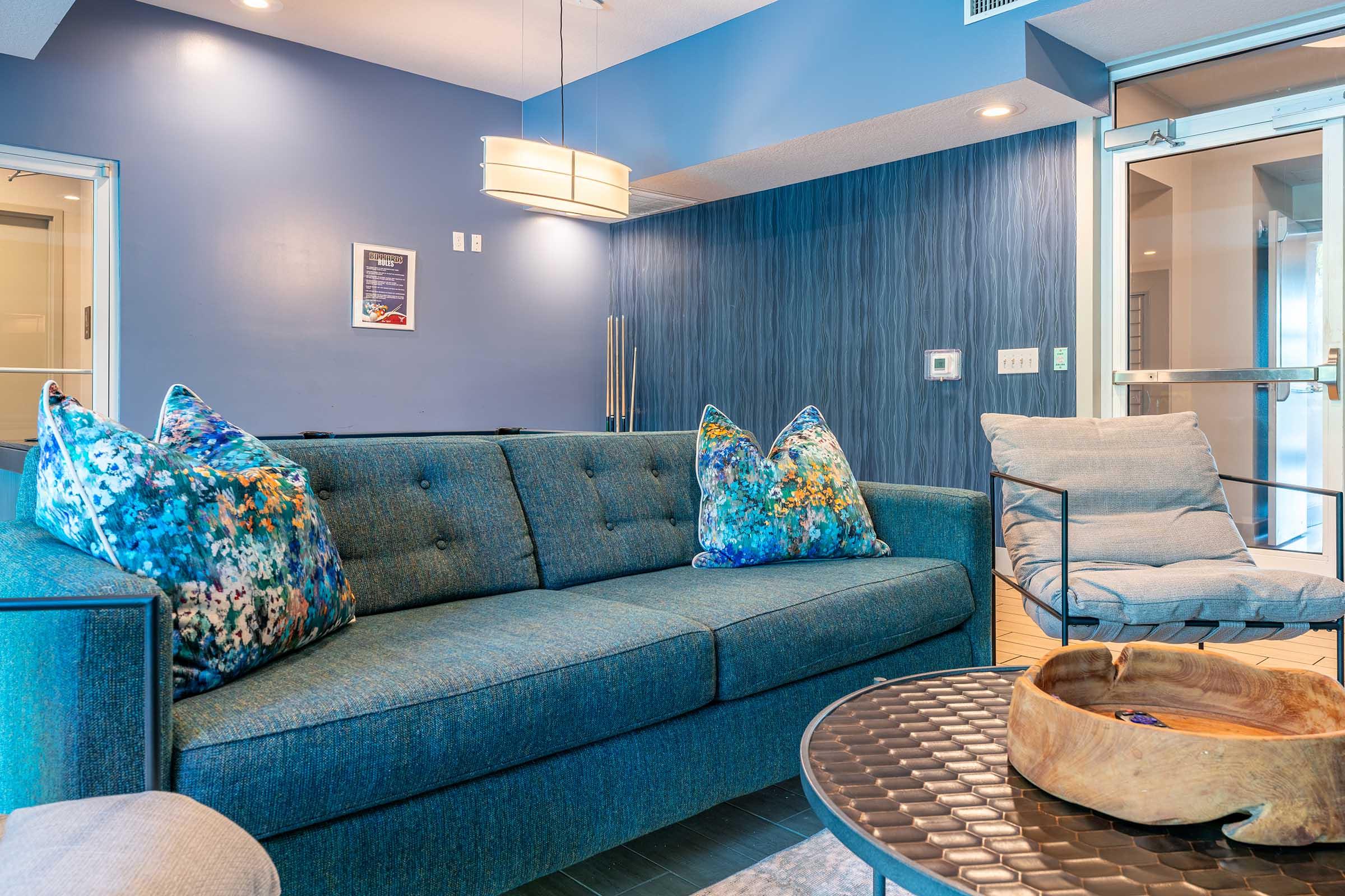 A modern living room featuring a blue sofa with colorful cushions, a decorative wooden bowl on a coffee table, and a wall with a blue textured design. Ambient lighting from a pendant fixture creates a cozy atmosphere, while a glass door leads to another space.