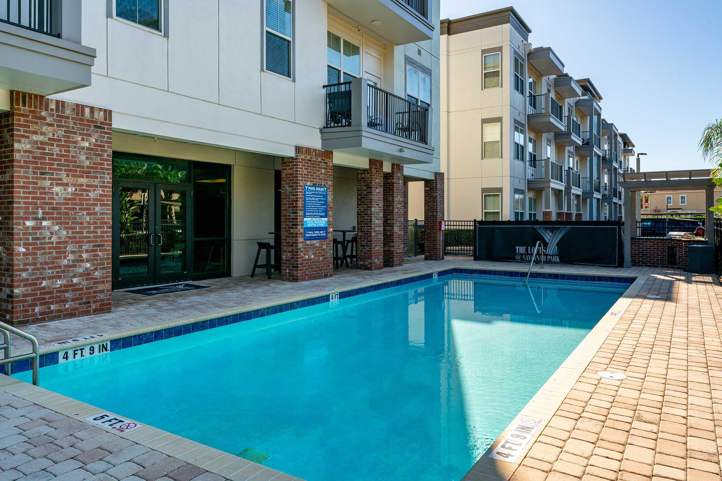 A clean, blue swimming pool surrounded by brick and stone walkways, with modern apartment buildings in the background. The area features a wall of windows, a poolside sign, and lounge chairs. Sunlight reflects off the water, creating a bright and inviting atmosphere.