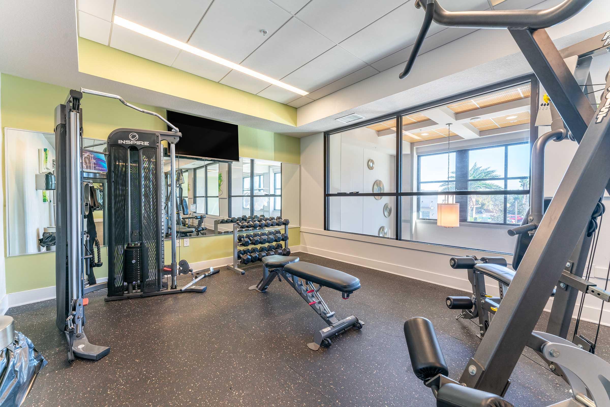 A modern gym interior featuring a variety of exercise equipment. A multi-station weight machine is visible, along with a set of dumbbells. There are benches and free weights, all situated in a well-lit space with large windows providing natural light. A mirror reflects the workout area, enhancing the open feel.