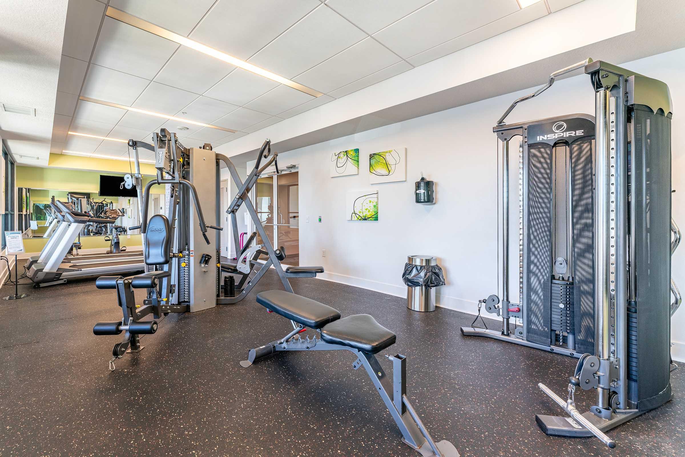 A modern gym interior featuring various workout equipment, including weight machines and benches. There are exercise bikes and treadmills in the background, along with bright green artwork on the walls and a trash can. The floor is covered with black rubber matting for safety and comfort.