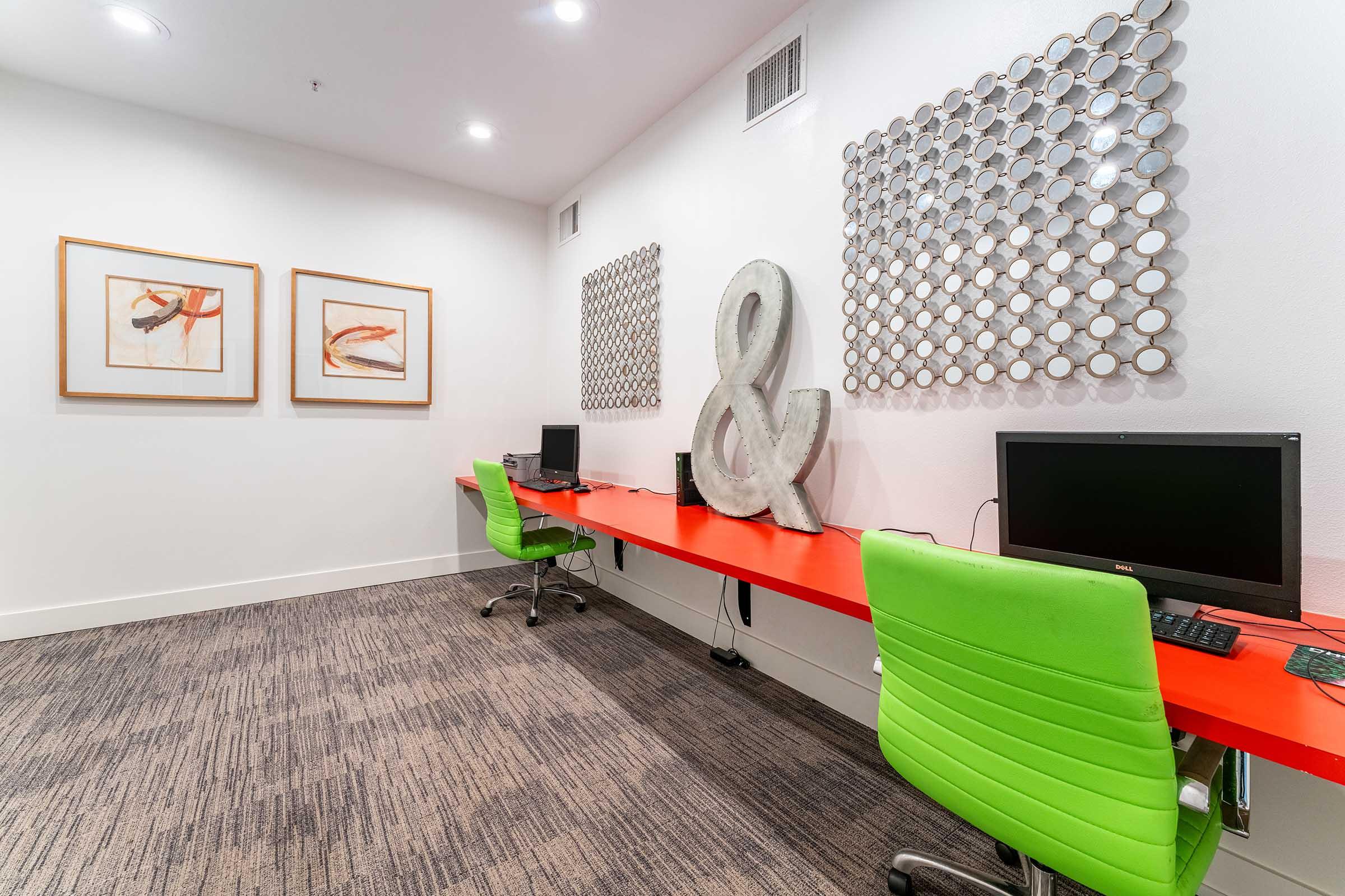 A modern workspace featuring two green office chairs at a long red desk with computers. The wall displays two framed art pieces and two decorative wall panels made of circular mirrors. The overall design is bright and contemporary, creating an inviting atmosphere for productivity.