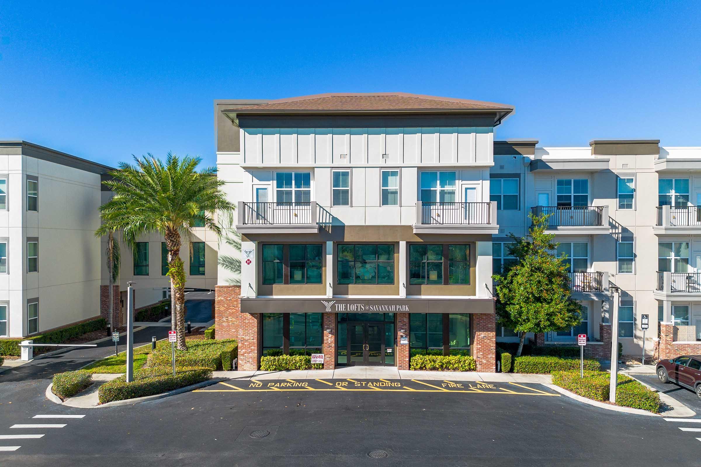 A modern multi-story building with a flat roof and balconies, surrounded by palm trees and landscaped shrubs. The entrance is marked with the name "The Hub's Savannah," and there are no-parking signs in front, indicating designated areas. The sky is clear and blue, creating a bright atmosphere.