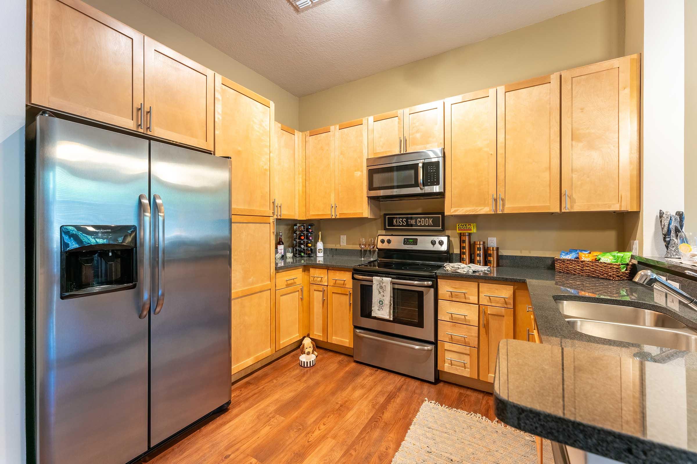 Modern kitchen featuring stainless steel appliances, including a refrigerator and oven, warm wooden cabinetry, and a dark countertop. The layout includes a sink and various kitchen items on the counter, along with a small decorative figurine on the floor. Natural light illuminates the space.