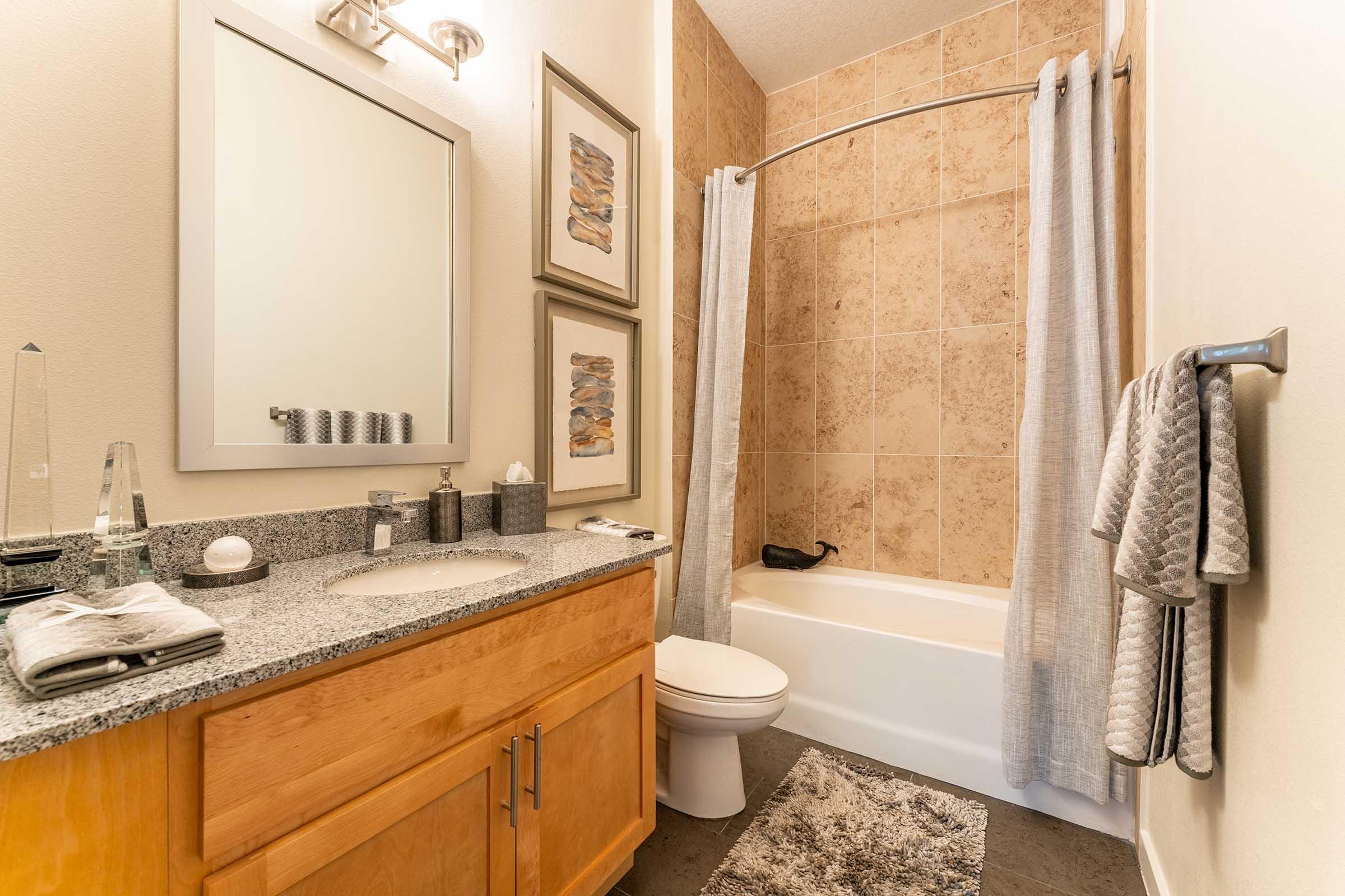 A modern bathroom featuring a clean design with beige tiled walls, a bathtub with a shower curtain, a simple wooden vanity with granite countertop, a mirror, and decorative wall art. The space is organized with towels and toiletries neatly arranged, creating a fresh and inviting atmosphere.