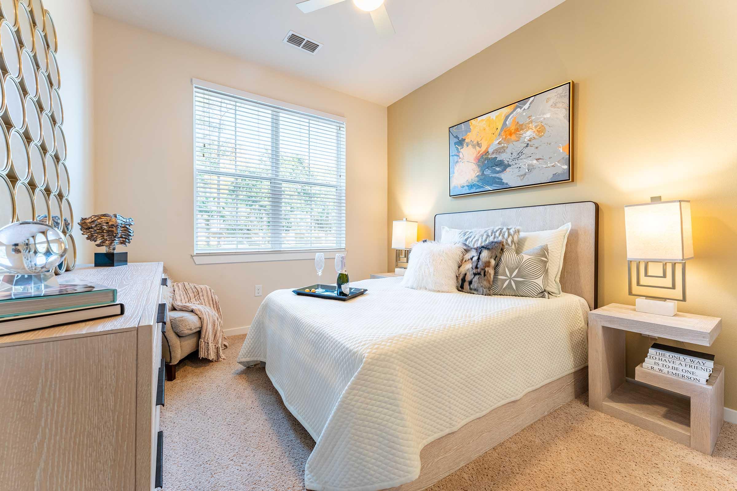 A cozy bedroom featuring a queen-size bed with a light quilt, decorative pillows, and a stylish headboard. A bedside table with a lamp is on each side, while a dresser with decorative items is visible. The room has light walls, a large window with natural light, and artwork on the wall.