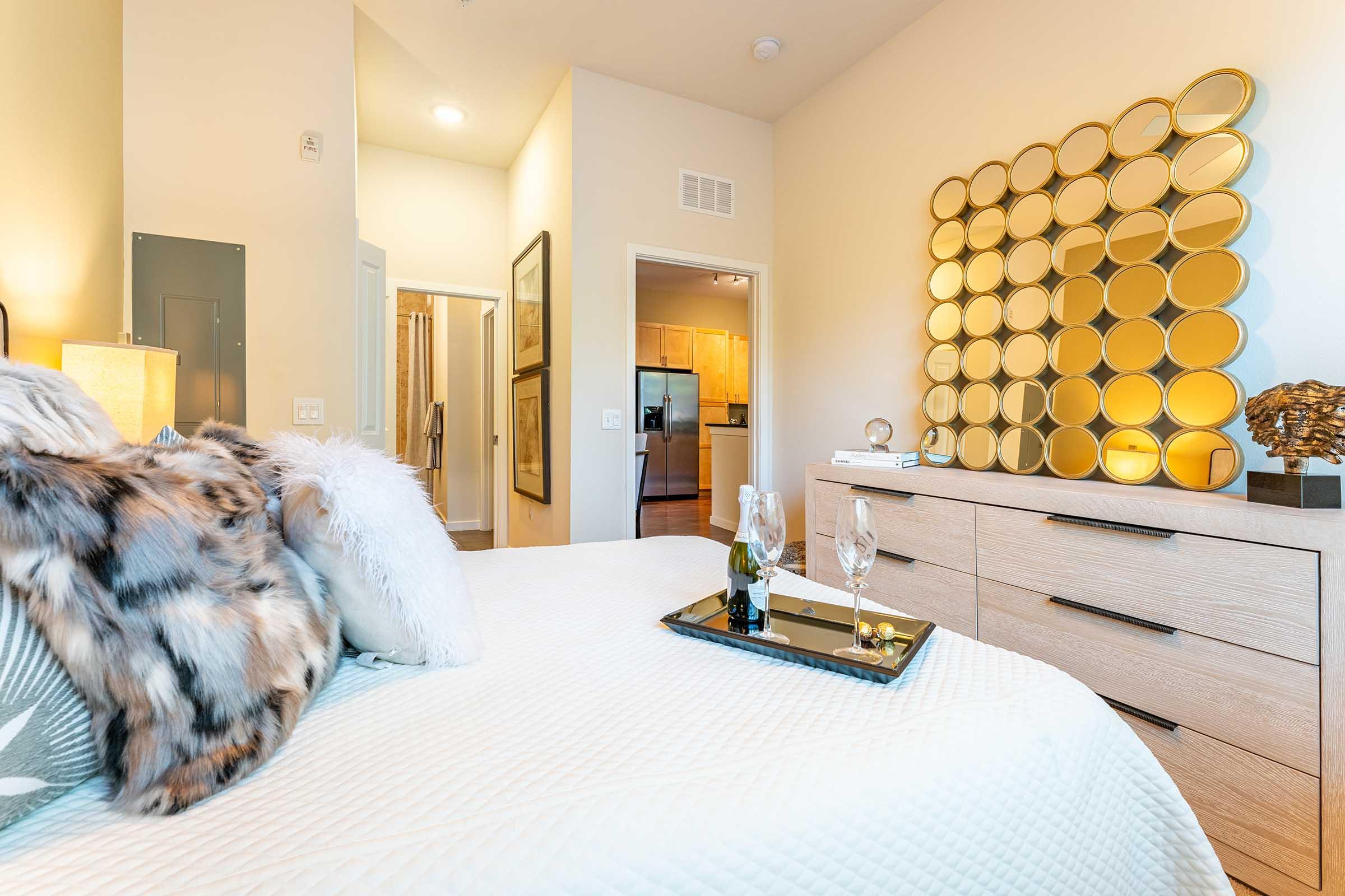 A cozy bedroom featuring a plush bed adorned with decorative pillows and a faux fur throw. A sleek dresser with a round mirror setup reflects elegance, while a tray holds two champagne glasses and a bottle, enhancing the luxurious ambiance. Soft lighting creates a warm, inviting atmosphere.