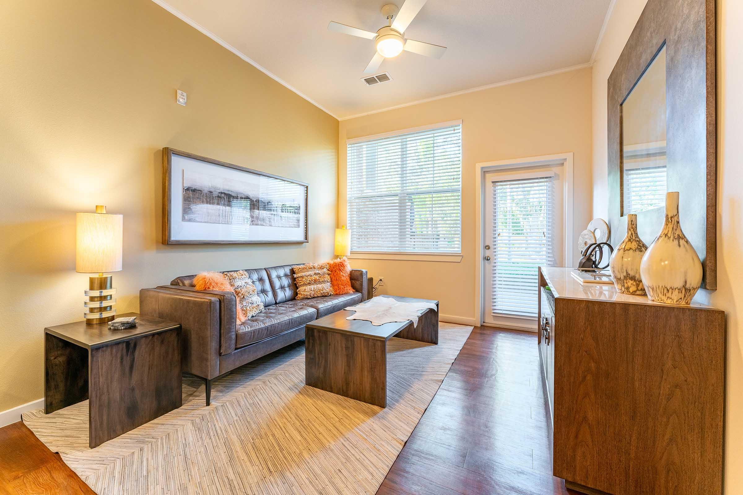 A cozy living room featuring a modern sofa with orange and white decorative pillows, a wooden coffee table, and a side table with a lamp. Natural light streams through large windows, illuminating the space. A sideboard with decorative vases and a mirror adds to the inviting atmosphere.