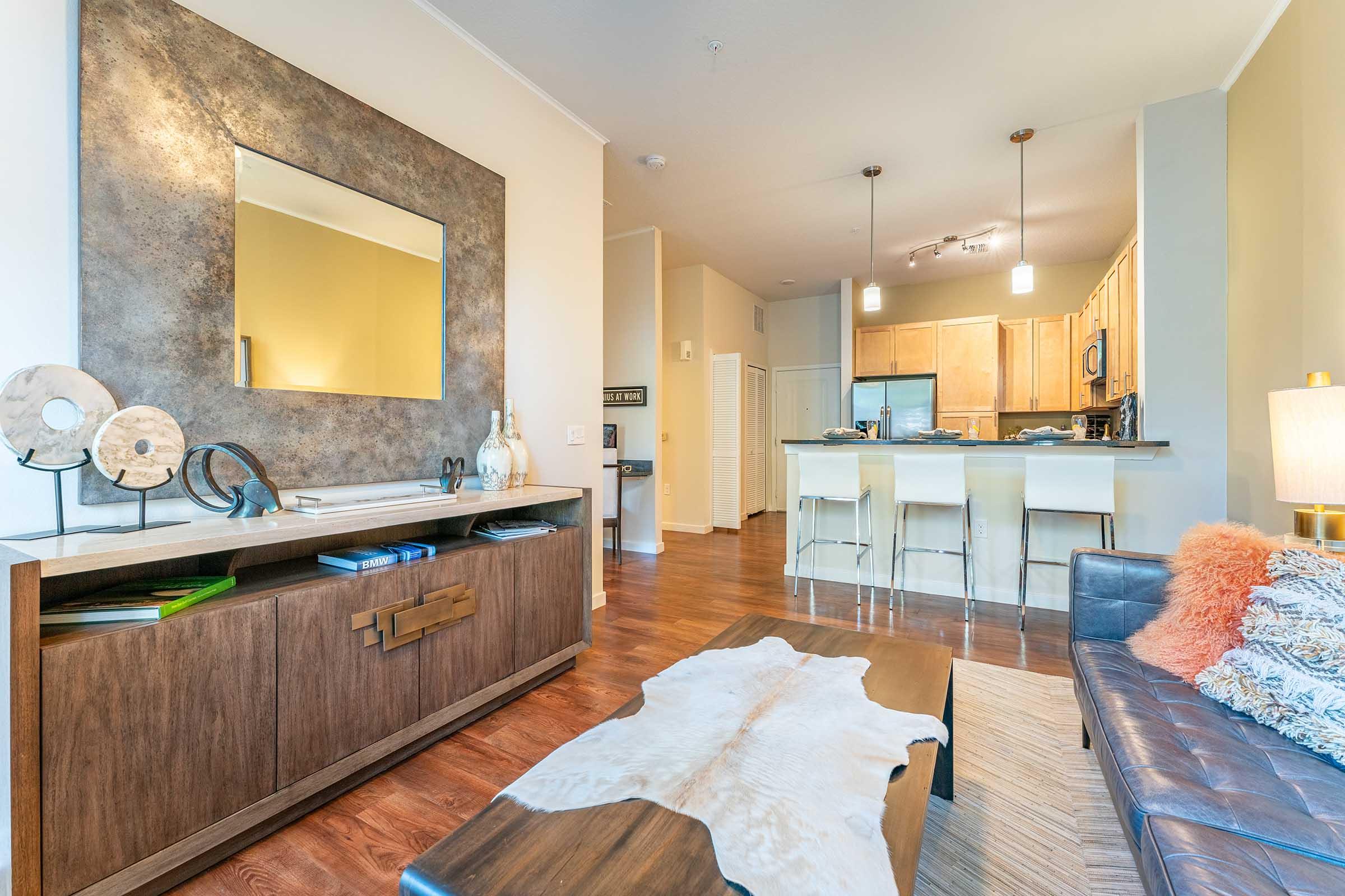 A modern living room featuring a sleek wooden coffee table with a decorative item on top, a brown leather sofa with colorful pillows, and a stylish mirror above a sideboard. An open kitchen with bar stools is visible in the background, accented by warm lighting and hardwood floors.