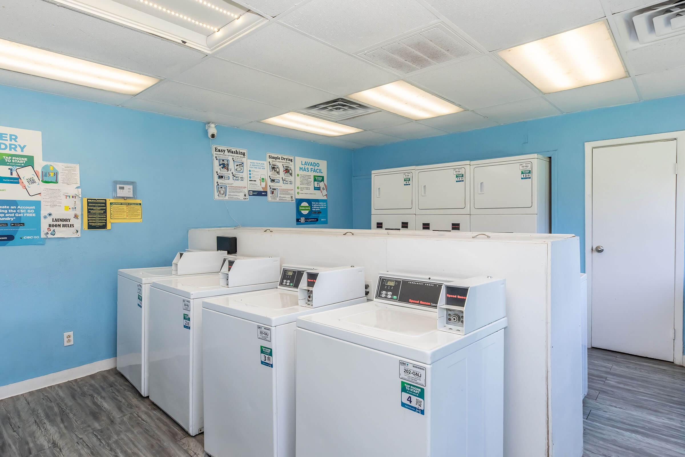 A clean, bright laundry room featuring several white washing machines and a row of white dryers against a blue wall. Informational posters about laundry services are displayed on the walls, and the space has a modern, well-maintained appearance with overhead lighting and a door on the right.