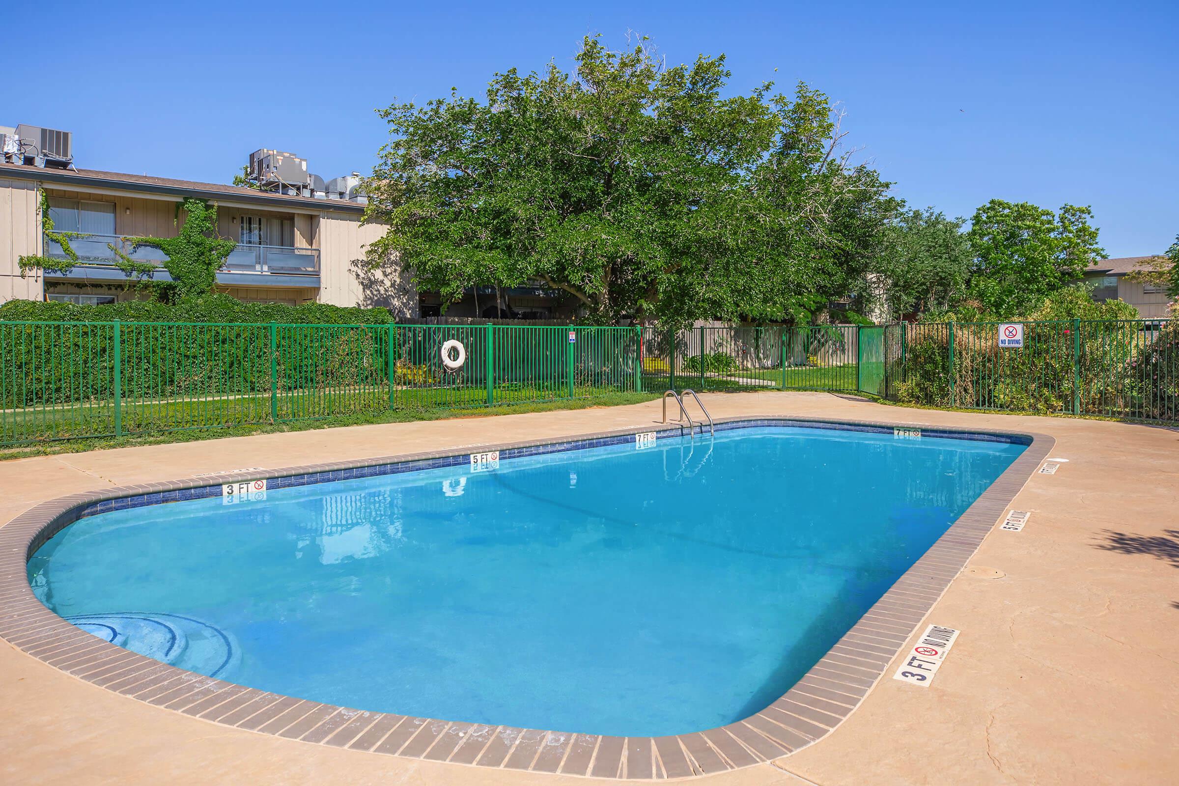 A clear swimming pool surrounded by a paved area and green landscaping. The pool has steps leading in and is enclosed by a green fence. In the background, there are trees and apartment buildings, suggesting a residential area. The sky is blue with no clouds visible.