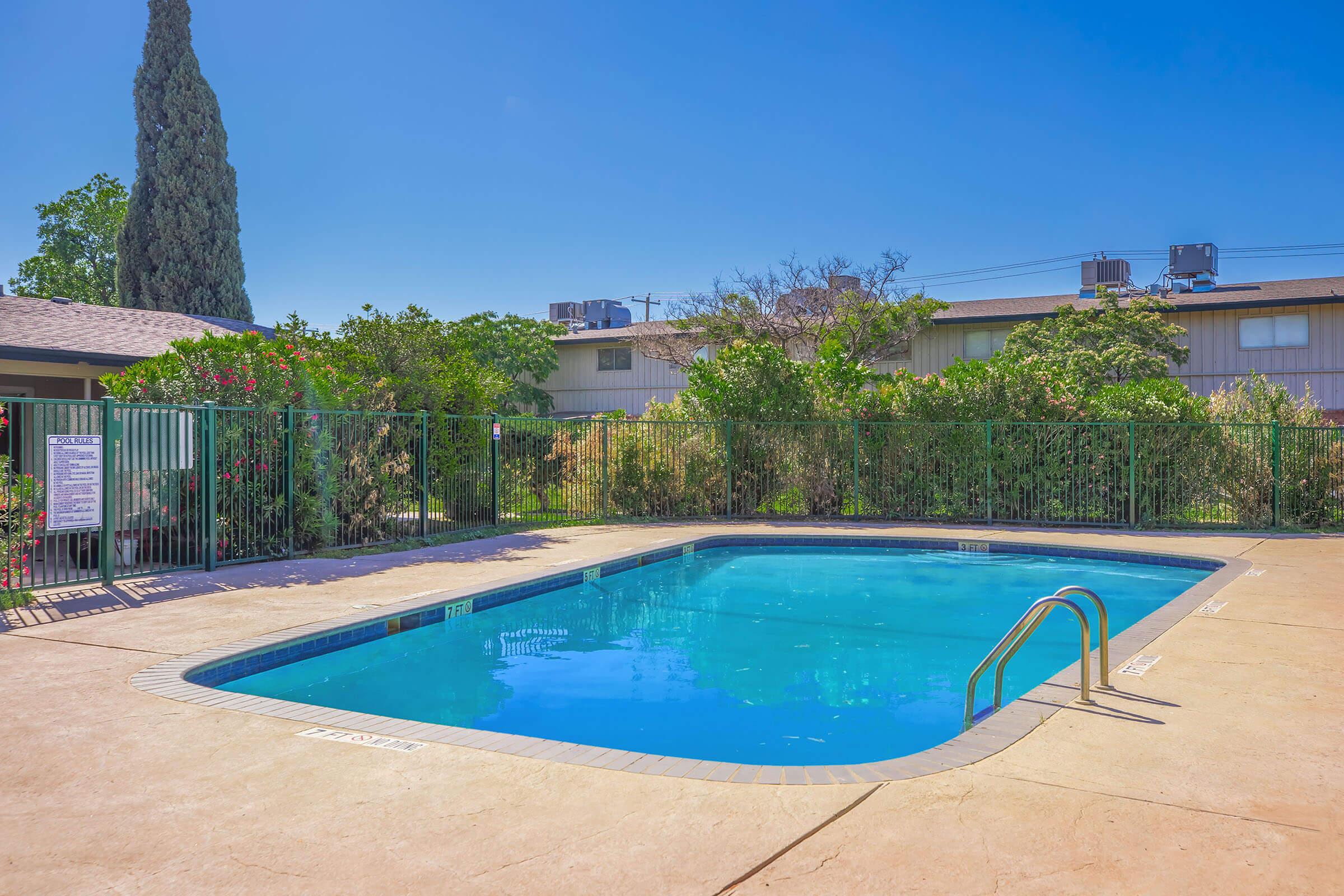 A clear blue swimming pool surrounded by a secure green fence. Lush greenery and flowering plants are visible nearby, with a residential building in the background. The sky is sunny and bright, creating a inviting outdoor atmosphere.