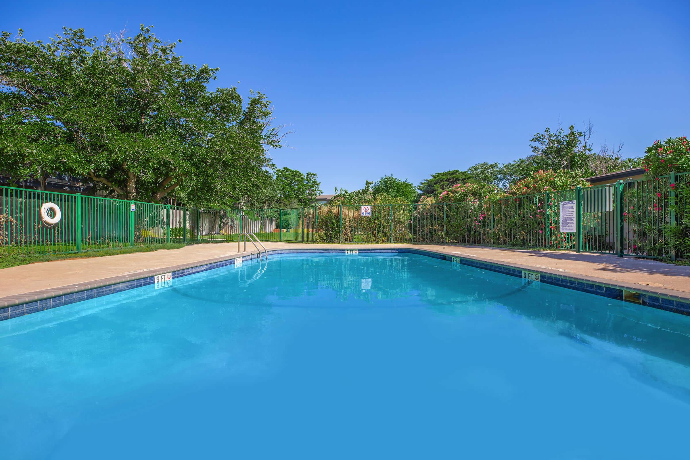 A clear blue swimming pool surrounded by a lush green landscape. The pool features a set of stairs leading into the water, with green fencing and trees in the background. Bright blue sky overhead enhances the tranquil and inviting atmosphere of the area.
