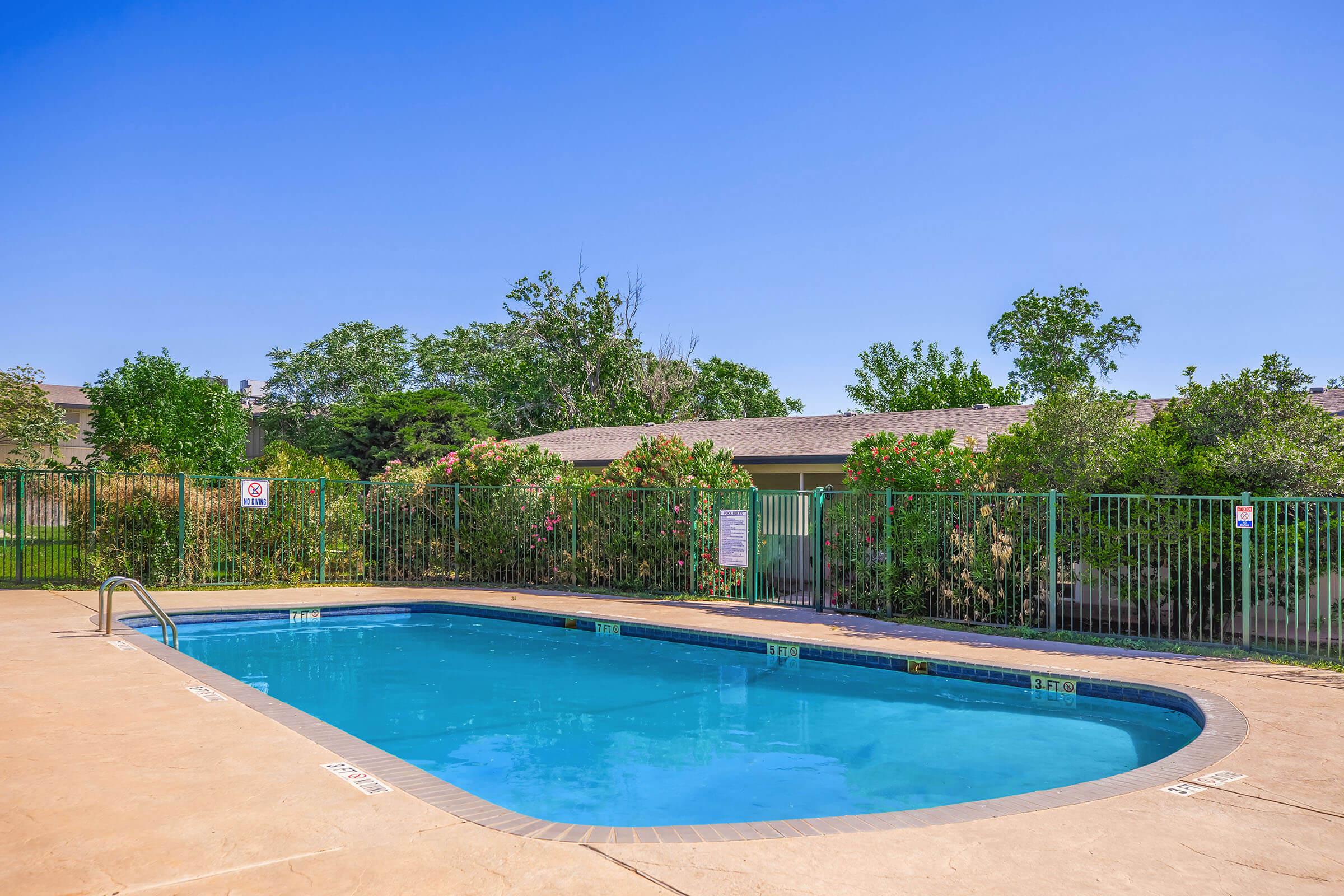 A clear blue swimming pool surrounded by green grass and shrubs, with a fenced area and a building in the background. The sky is bright and sunny, enhancing the pool's inviting appearance.