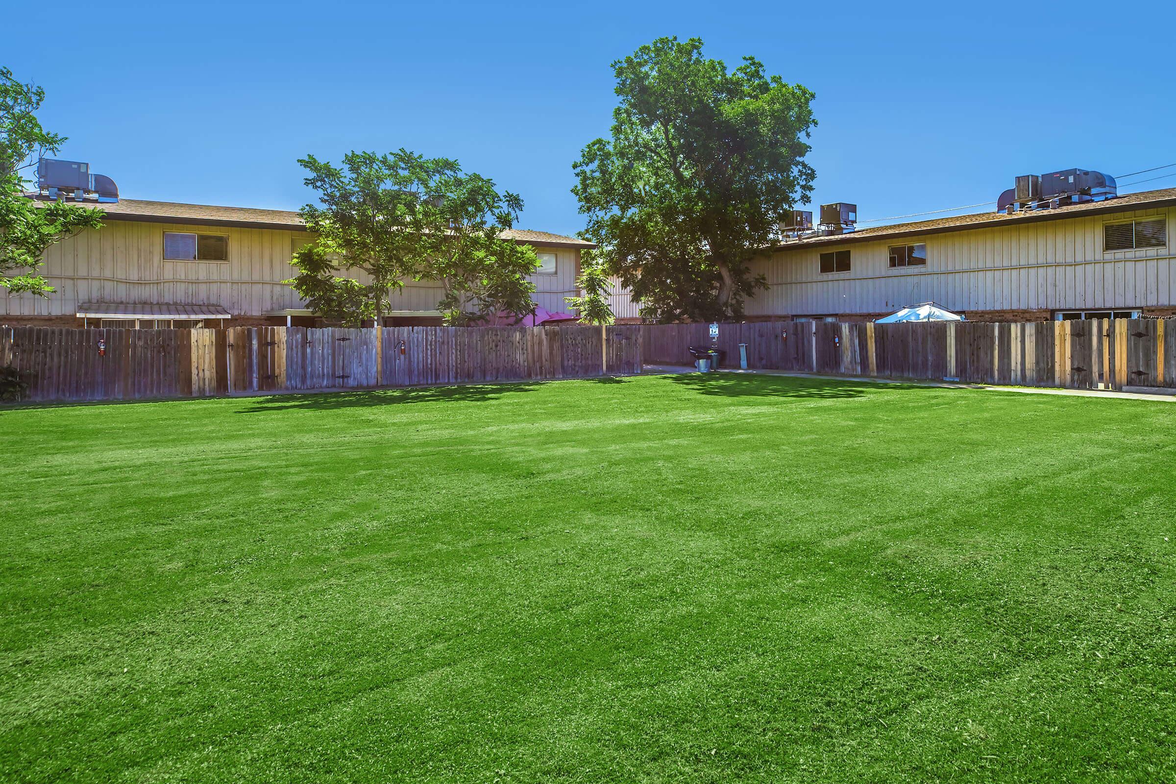 A spacious green lawn enclosed by wooden fences, with a few trees and two apartment buildings in the background. The sky is clear blue, and there are air conditioning units on the rooftops of the buildings. The scene conveys a bright, inviting outdoor space.