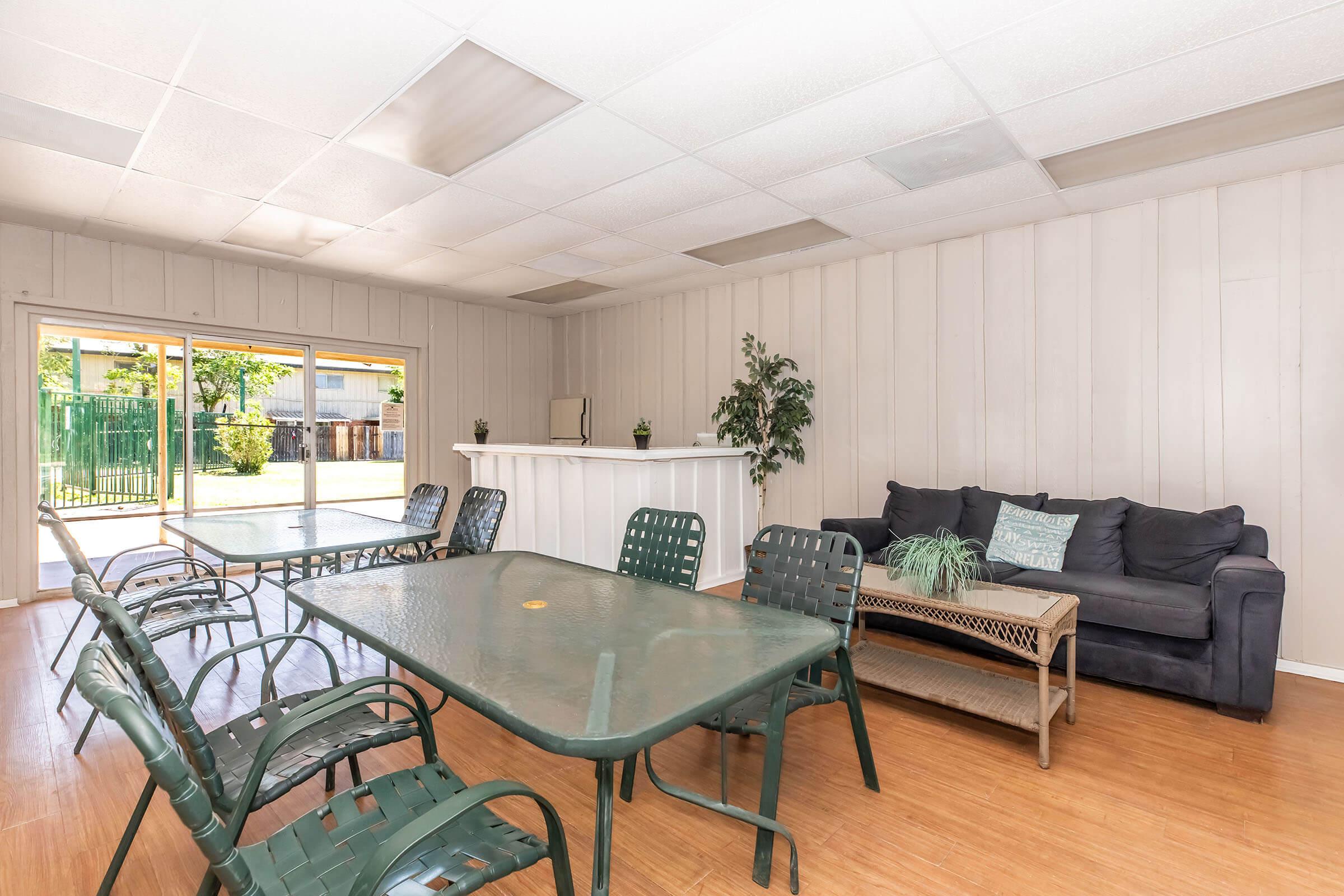 A spacious, well-lit room featuring multiple green metal tables surrounded by chairs. A comfortable dark sofa with decorative pillows is positioned against the wall. Large windows let in natural light, and there's a bar-like counter at the far end of the room. Greenery is visible outside through the windows.