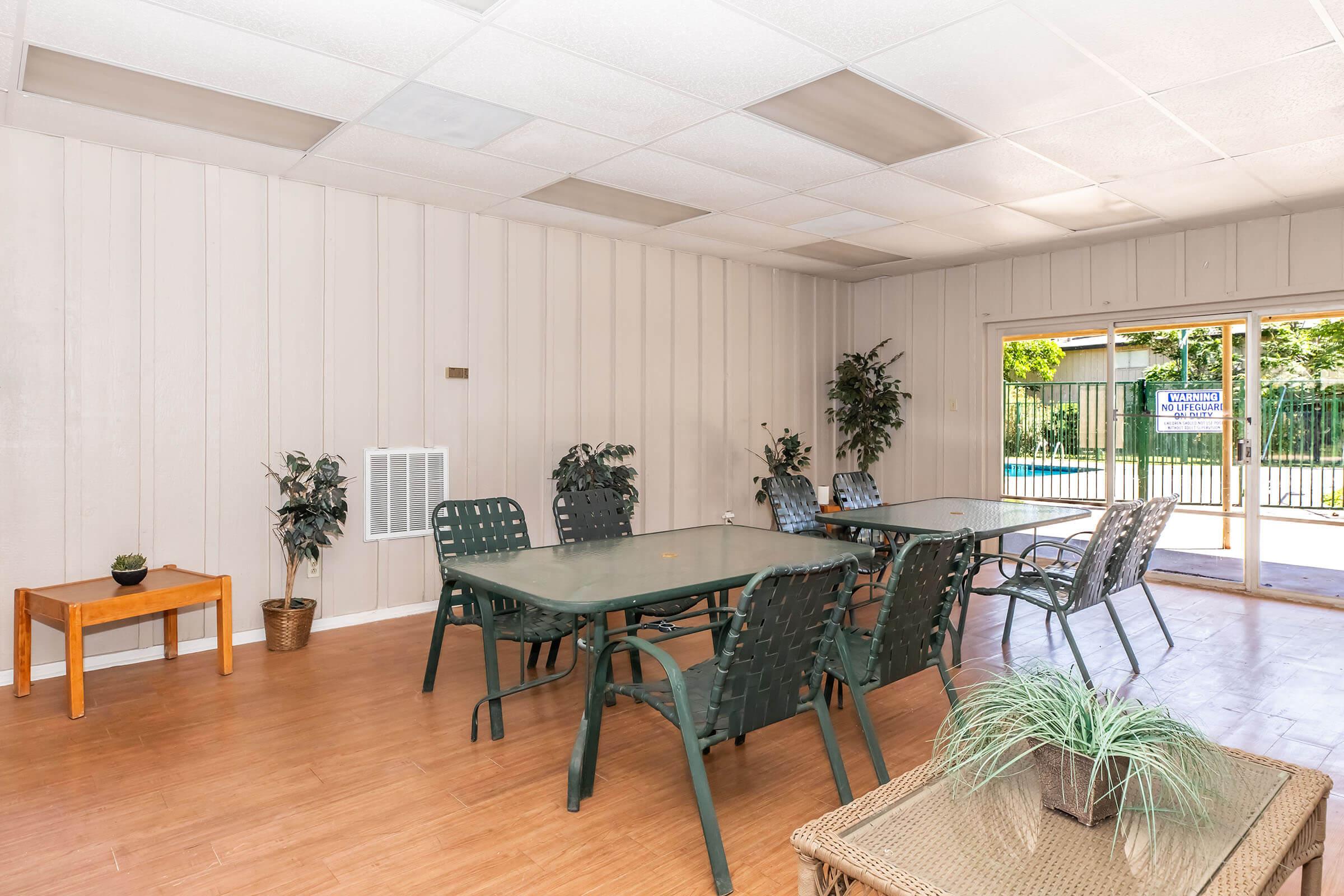 A spacious indoor area featuring several glass-top tables with green chairs, potted plants, and a wooden table. Large windows provide a view of an outdoor space, which includes a swimming pool and a fenced yard. The walls are light-colored, and the ceiling has grid tiles.