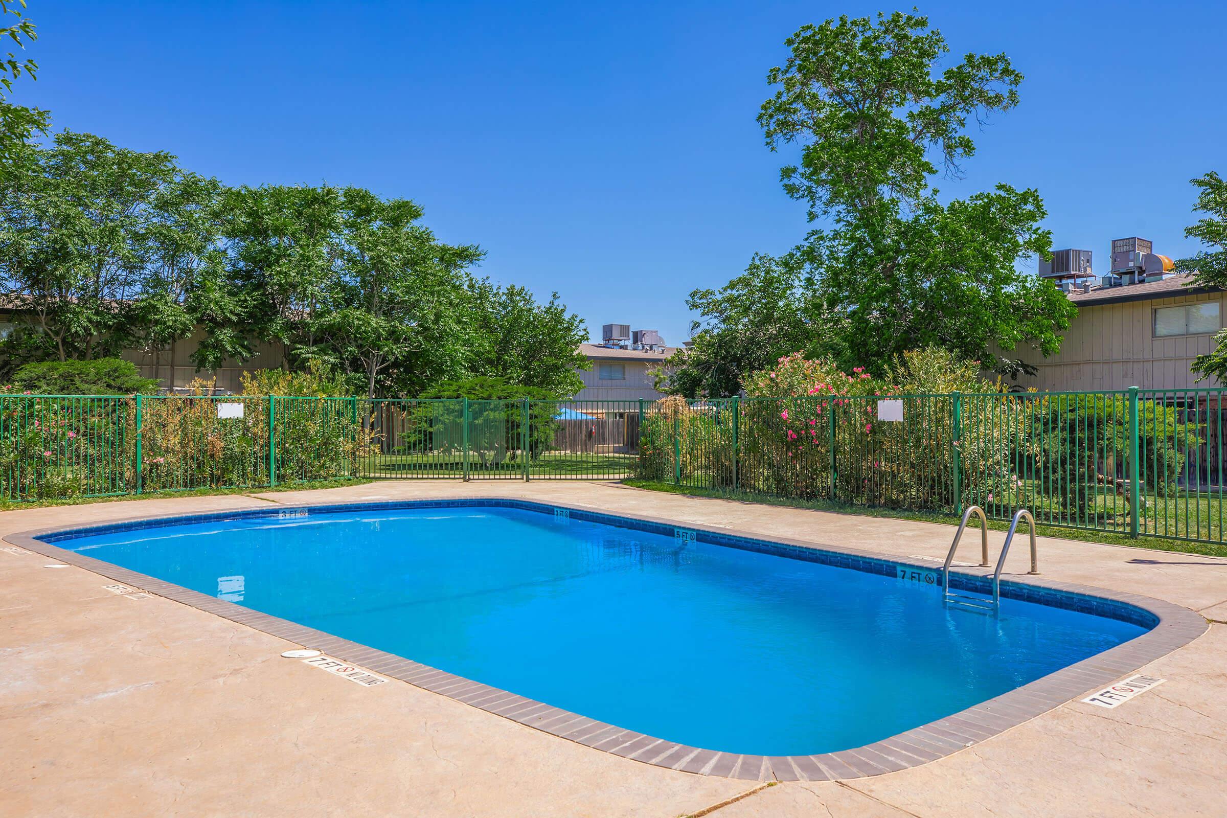 A clear, inviting swimming pool surrounded by a concrete deck and green fencing. Lush greenery and bushes with blooming flowers are visible nearby, under a bright blue sky. The pool area appears well-maintained and is suitable for relaxation and recreation.