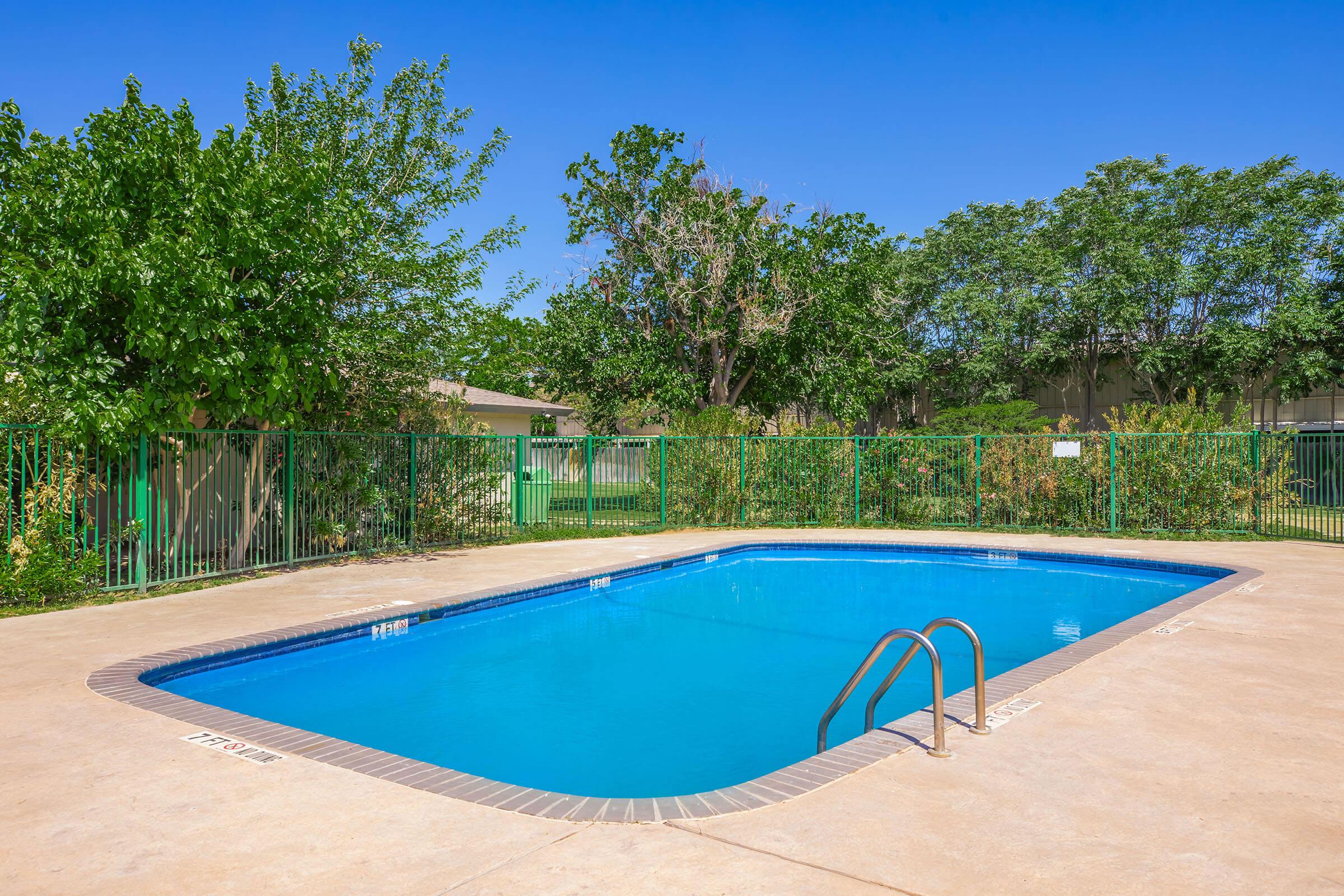 A clear blue swimming pool surrounded by a tan concrete deck. Lush green trees and shrubs are visible in the background, alongside a green fence enclosing the pool area. The sky is bright blue with no clouds, creating a sunny and inviting atmosphere.